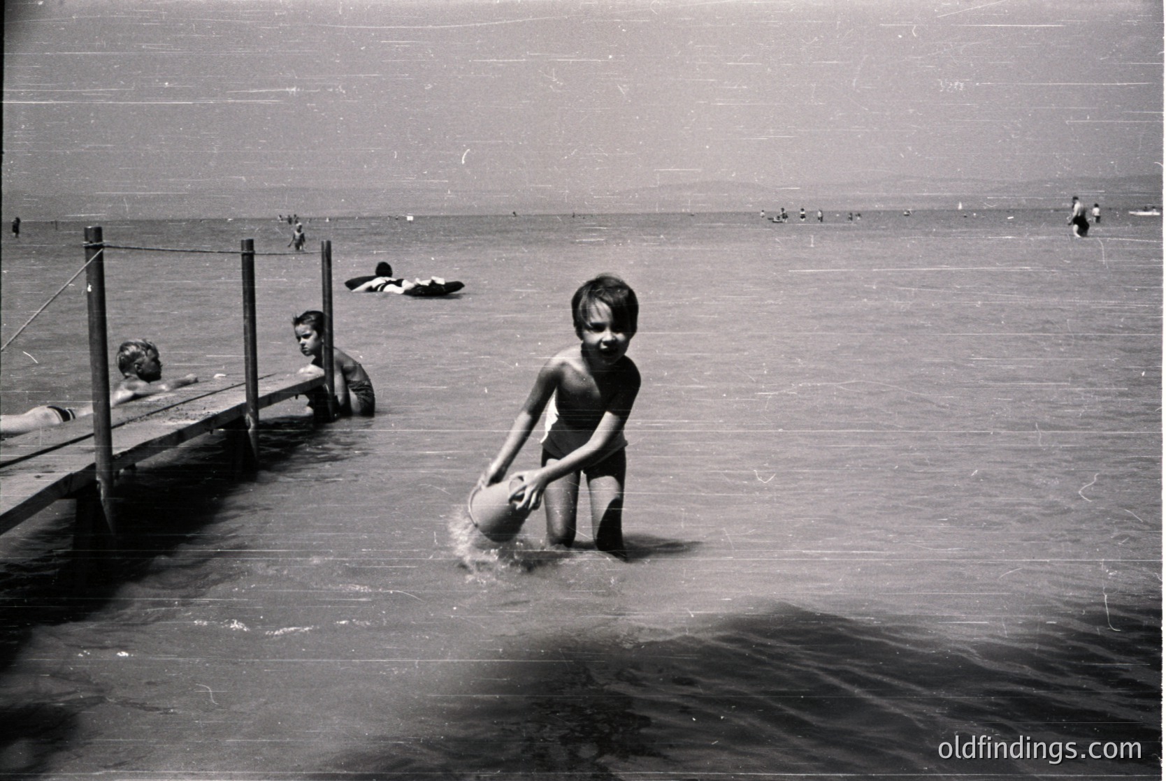 Mid-20th century beach scene with wooden pier and shallow waters. A young boy in swim trunks holds a bucket, wading near the shore. Other swimmers and sunbathers visible in background. Classic vintage coastal lifestyle.