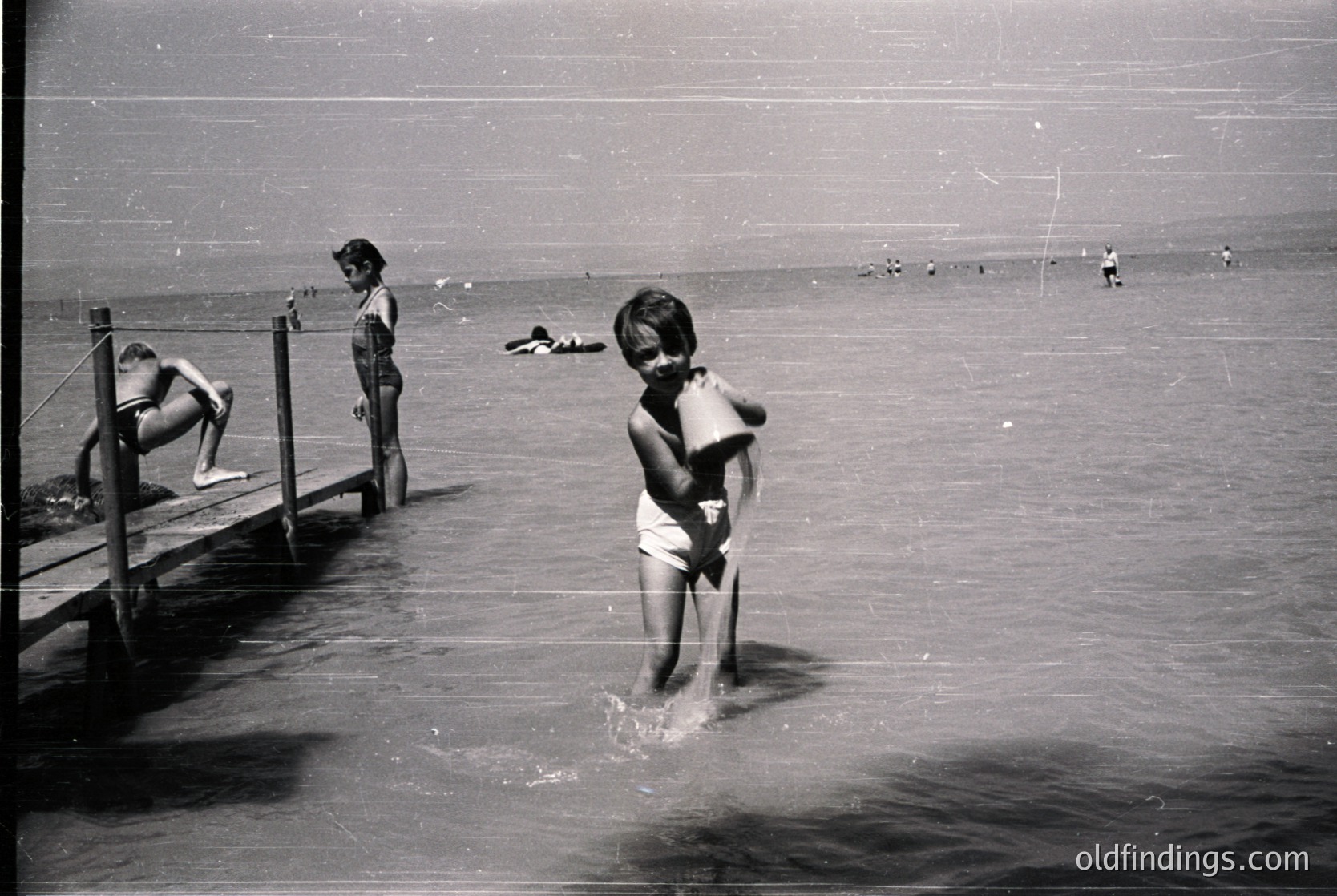 Mid-20th century seaside scene: children wade in shallow water near a wooden pier. Foreground boy in swim trunks clutches a towel; others sit/stand on pier. Crowded beach with distant swimmers.