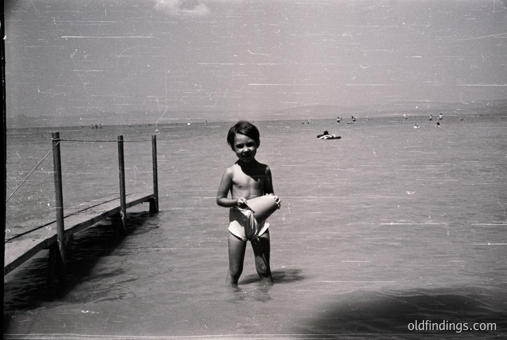 Young boy in mid-20th-century swim trunks stands waist-deep in shallow coastal waters, clutching a white cloth or towel. Wooden pier and railing frame left side; distant swimmers dot the horizon. Vintage black-and-white grain highlights 1950s-60s seaside nostalgia.