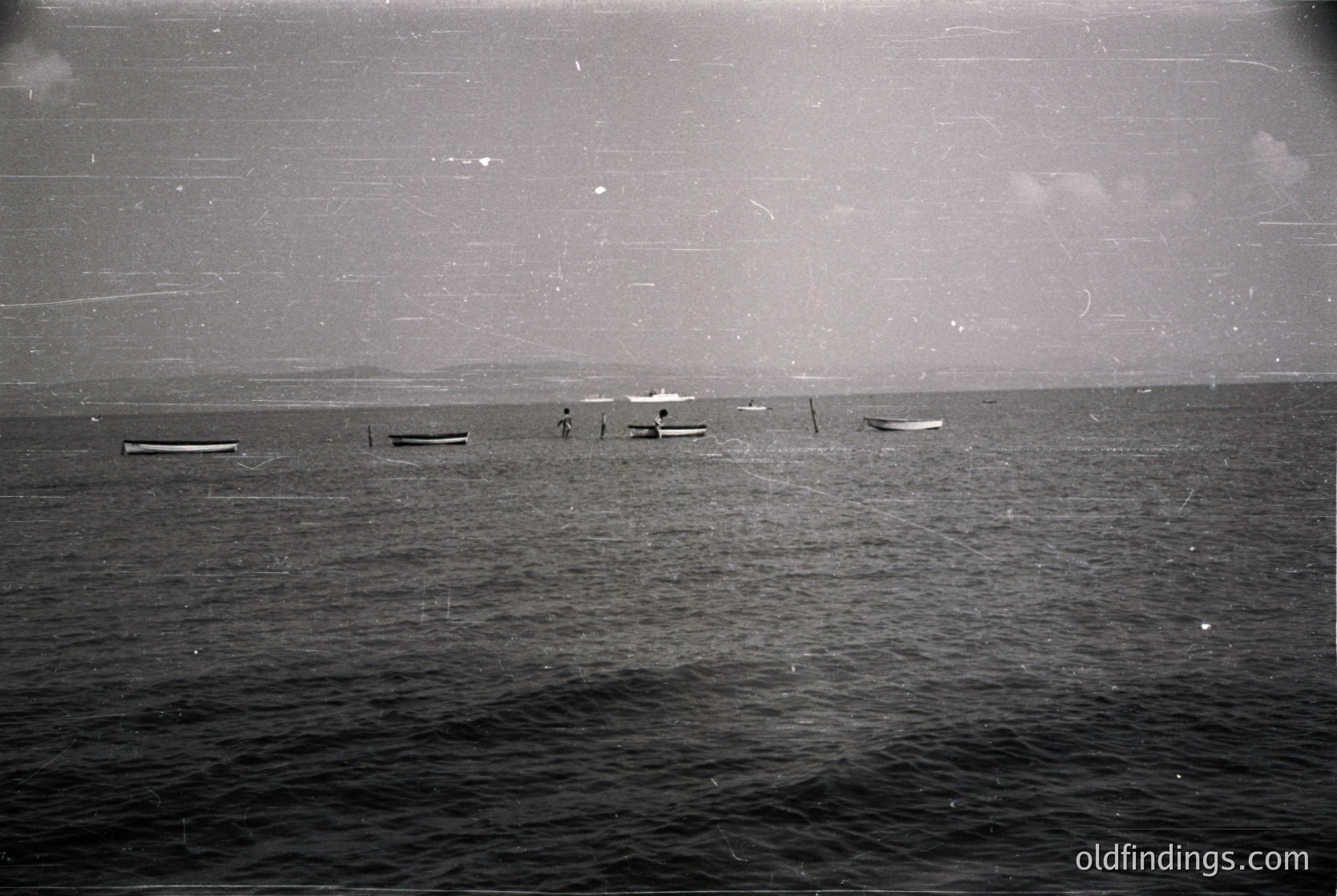 Vintage black-and-white seascape showing three small wooden boats anchored near shore, likely fishing vessels. Distant shoreline with faint outlines of landforms. Water appears calm with subtle ripples. Mid-20th century coastal scene, possibly Mediterranean or Black Sea region.