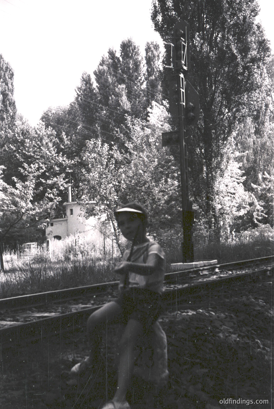 Mid-20th century black-and-white photo: man in uniform (likely military or railway worker) perched atop a train rail, holding a long pole. Dense forest and a small white building with a chimney in background. Rustic railway tracks and telegraph poles suggest rural or industrial setting.