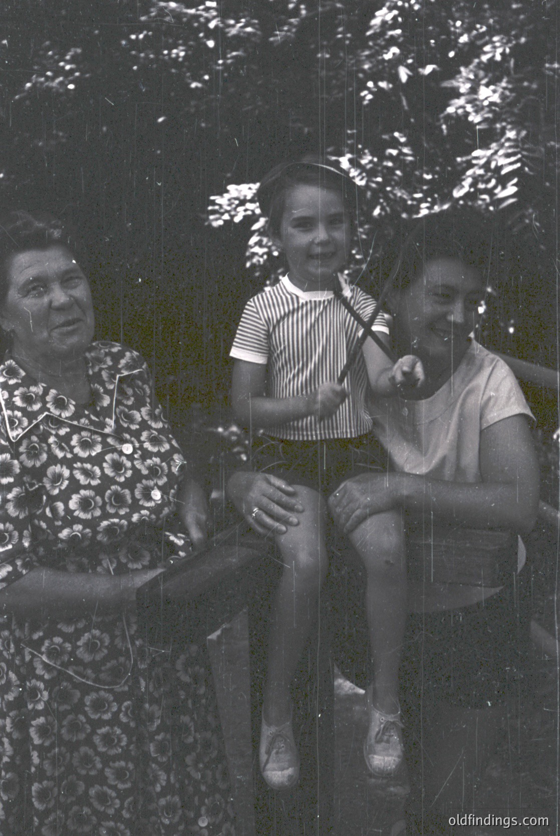 Three generations share a moment on a wooden swing in a lush, shaded park. Elderly woman in floral dress, striped shirt child, and younger woman in sleeveless top. Mid-20th century family portrait, likely 1950s–1960s.