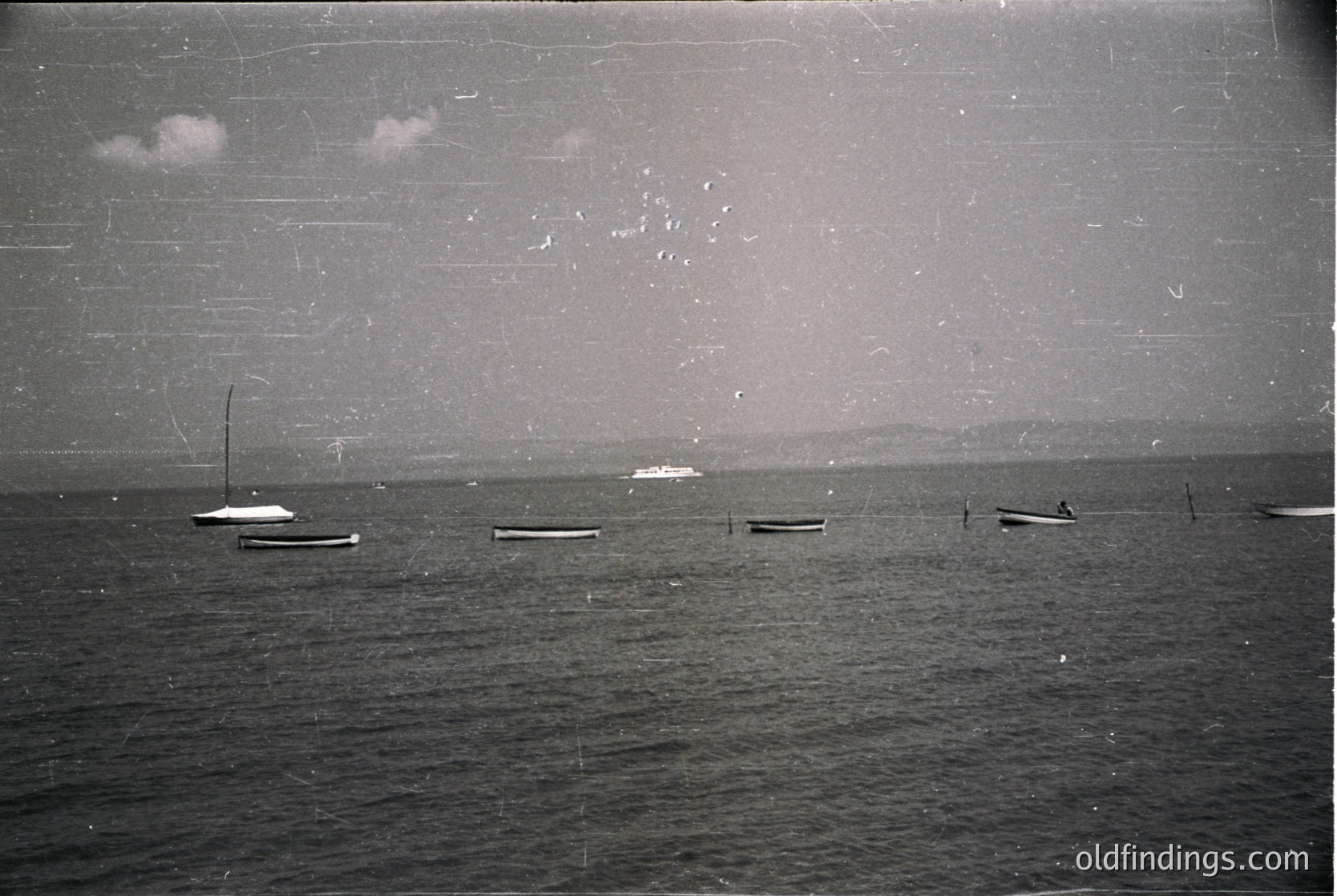 Vintage black-and-white seascape featuring small wooden fishing boats anchored in calm waters. Distant shoreline with faint industrial structures visible. Likely mid-20th century coastal scene, possibly Eastern European or Mediterranean.