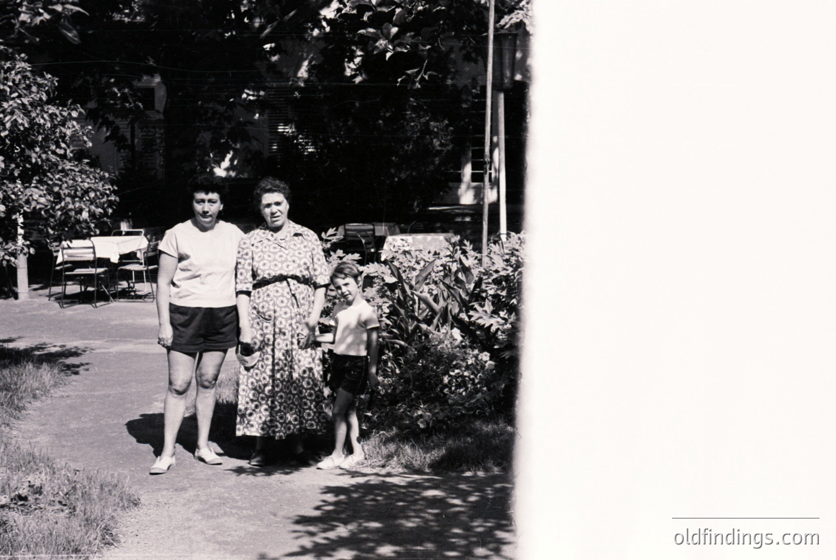 Black-and-white family portrait in a residential garden, mid-20th century. Adults in casual summer attire—man in short-sleeve shirt/shorts, woman in floral dress—stand beside a young boy in shorts. Lush greenery, wooden fence, and a parked car in background suggest suburban life. Authentic vintage aesthetic ideal for historical research or nostalgic design.