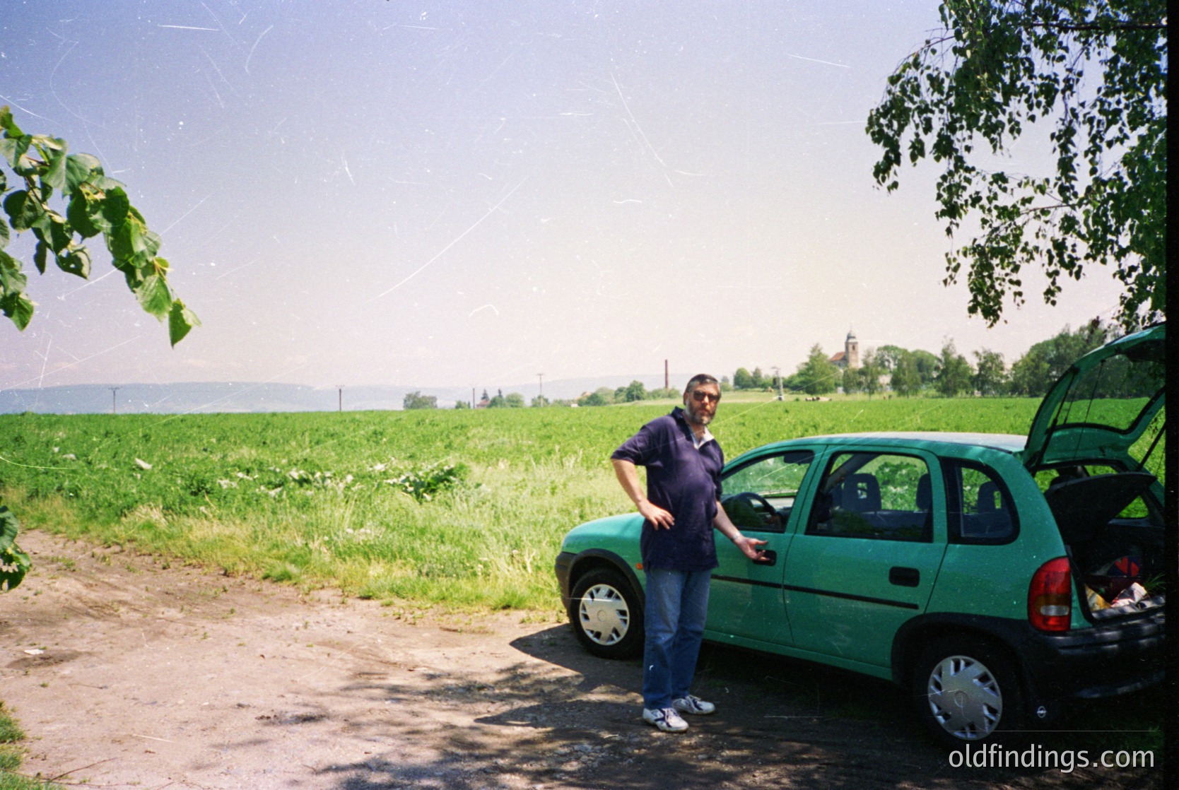 Vintage green hatchback (likely a Škoda Fabia or similar) parked beside a rural road, with a man in a dark polo shirt and jeans posing. Open trunk reveals minimal cargo. Lush green fields and distant trees suggest a countryside setting. Overcast sky with visible spiderwebs. Likely late 1990s–early 2000s.