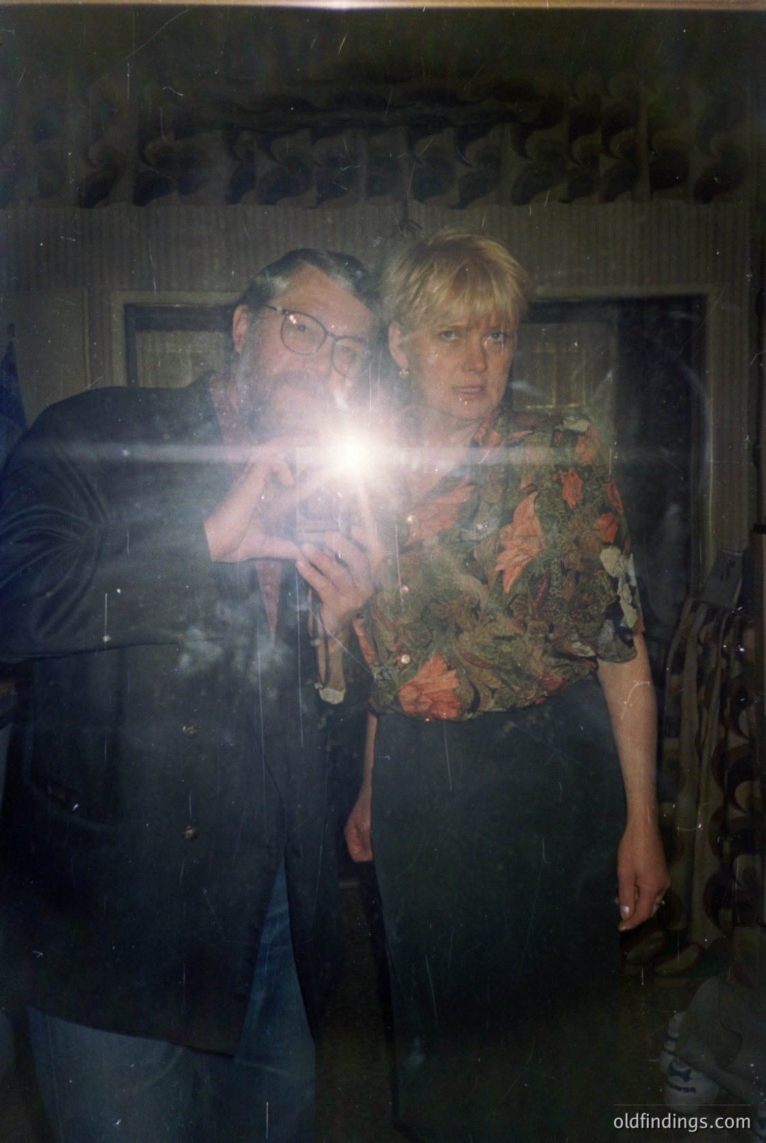 Vintage portrait of two individuals posing indoors, likely 1970s–1980s. Man in dark jacket, round glasses, and patterned tie; woman in floral blouse with high-waisted trousers. Warm lighting and blurred background suggest a candid, informal setting. Potential for nostalgic or fashion reference.