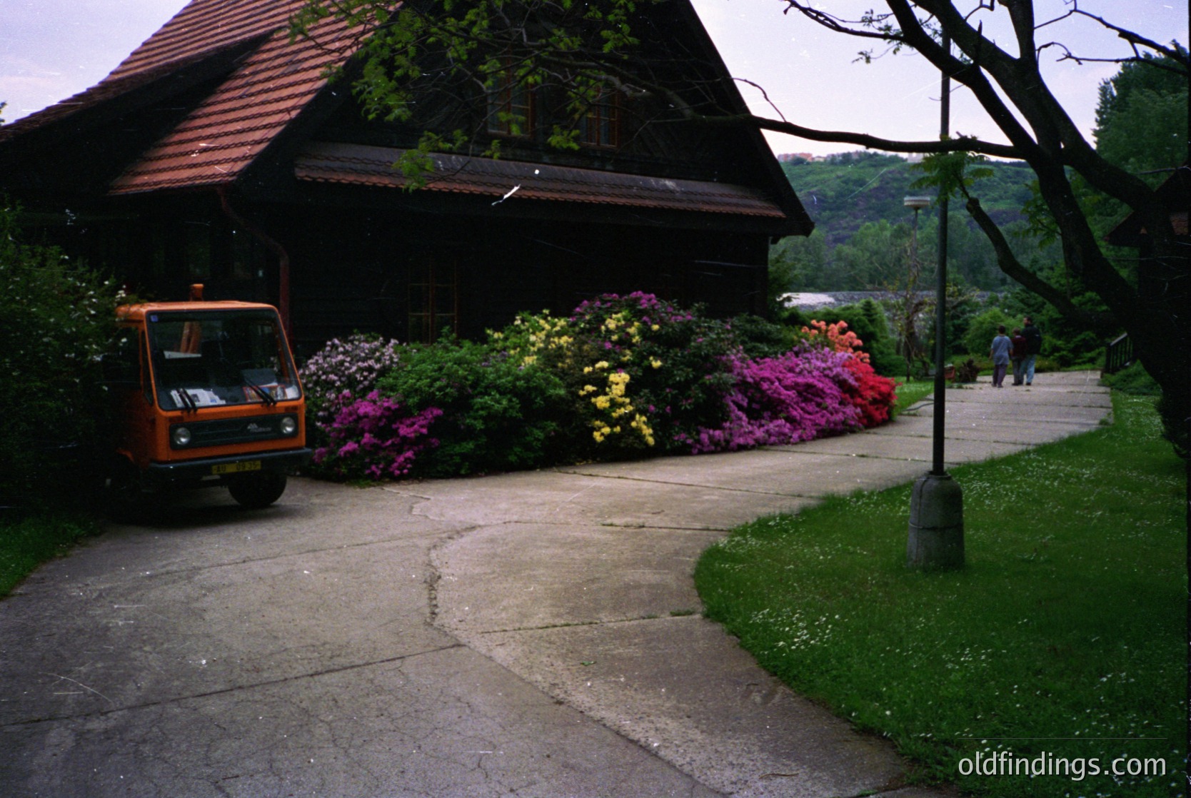 Vintage orange utility vehicle parked beside a lush garden bed bursting with vibrant pink, yellow, and purple flowers. Wooden chalet-style building with a sloped roof and overgrown greenery in the background. Pathway bordered by manicured grass leads toward a distant lake and forested hillside. Likely a mid-20th century European countryside setting.