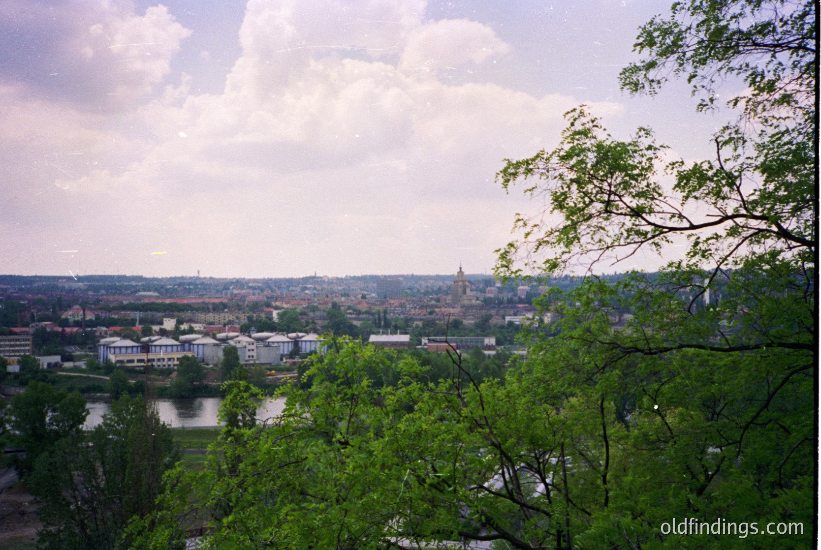 Panoramic view of a mid-20th century European cityscape with a river winding through urban and green zones. Distinctive church spire and low-rise residential blocks dominate the skyline. Lush foliage frames the foreground, suggesting a park or elevated vantage point.