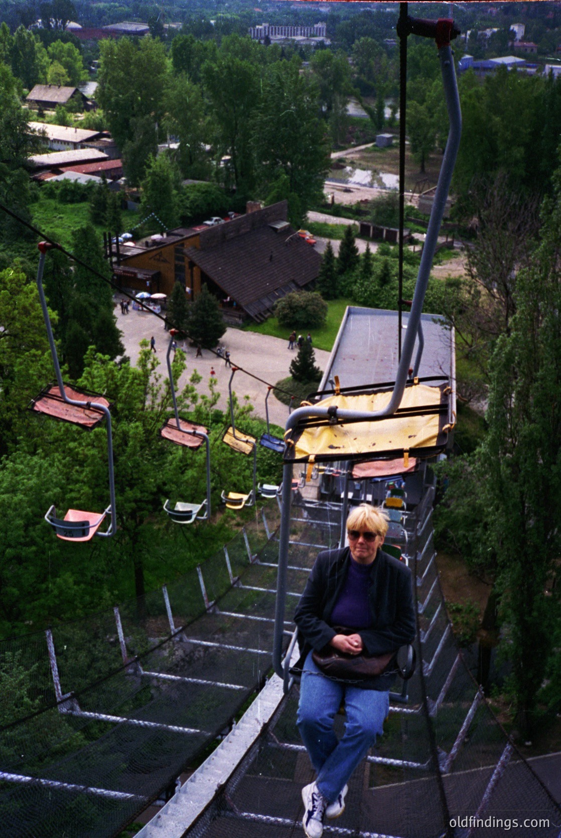A woman sits mid-ride on a vintage alpine cable car, surrounded by lush greenery and wooden cabins. The scene suggests a mid-20th century European mountain resort, likely or . The rustic design and cable infrastructure offer historical charm for tourism or design references.
