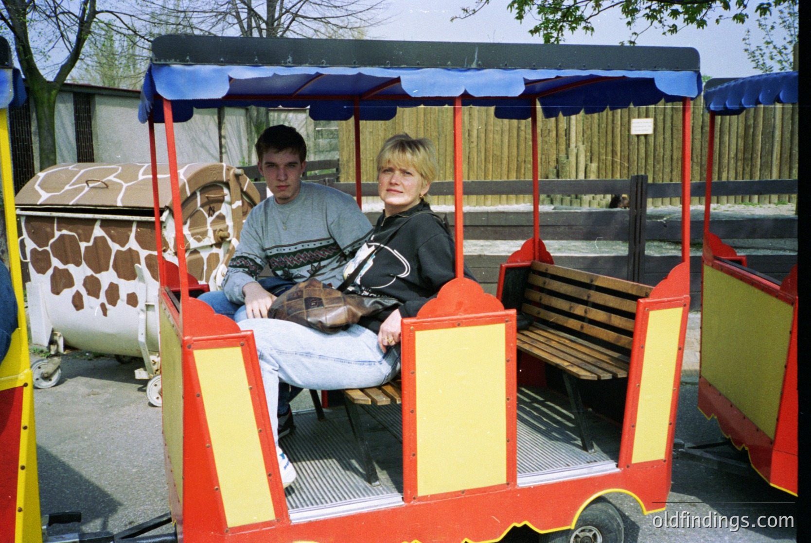Vintage zoo tram with bright red/blue canopy carrying two passengers in 1980s-90s attire. Giraffe enclosure with patterned skin mural visible in background. Casual, mid-century zoo experience.