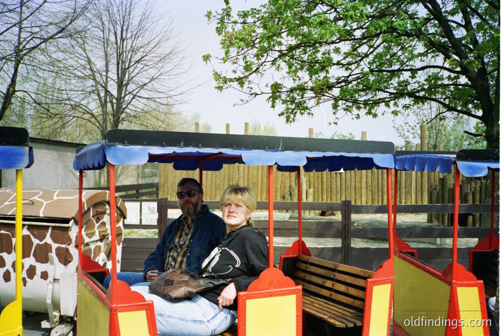 Brightly painted safari-style train cart with yellow, red, and blue accents, featuring a giraffe-patterned backdrop. Two adults seated inside, likely mid-1990s to 2000s. Indoor/covered zoo or wildlife park setting with wooden fencing and trees.