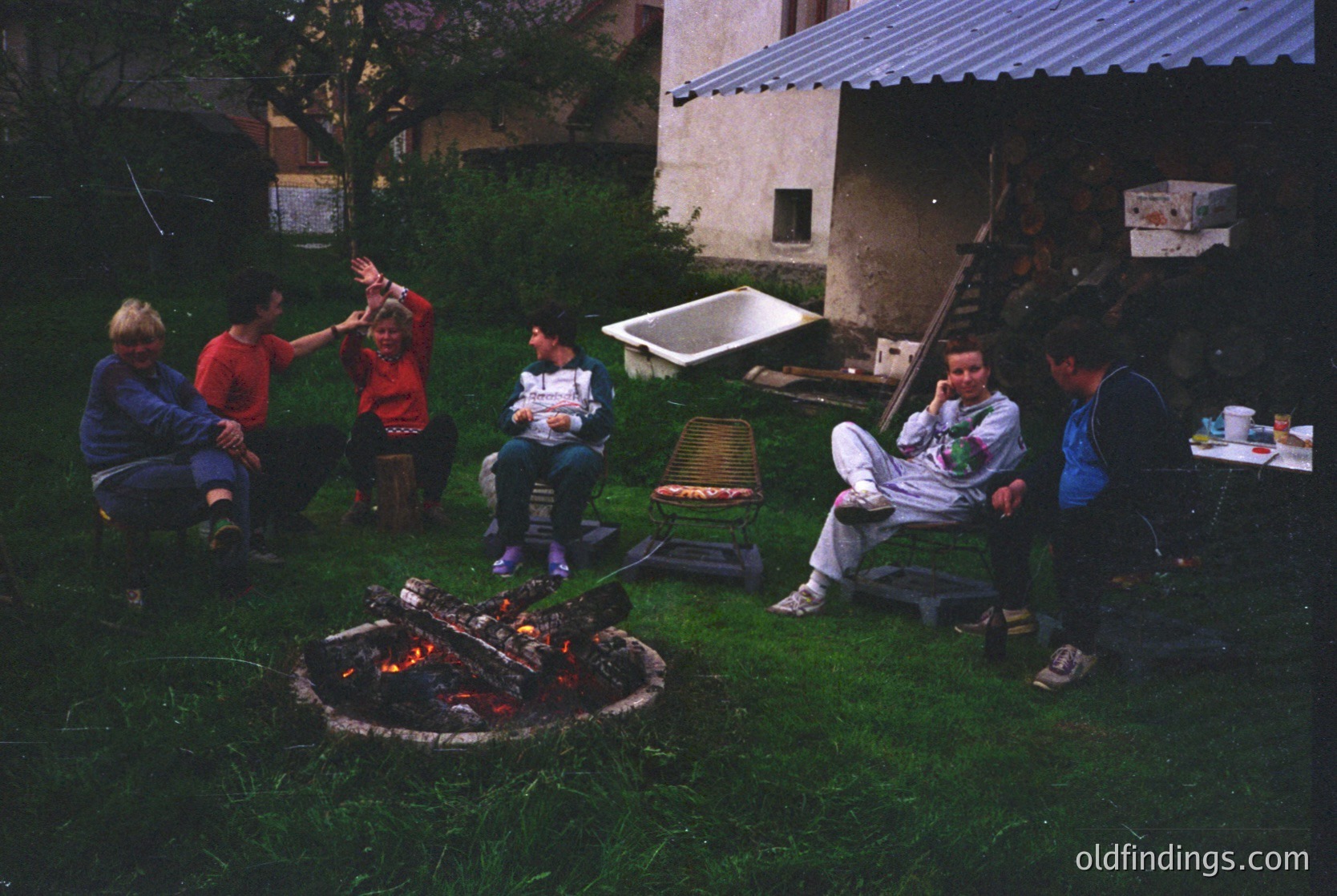 Family gathering around an outdoor fire pit in a residential courtyard, likely 1990s. Six adults and children seated on stools and grass, engaging in casual conversation. Concrete building with metal roofing and greenery in background. Authentic snapshot of communal backyard activities.