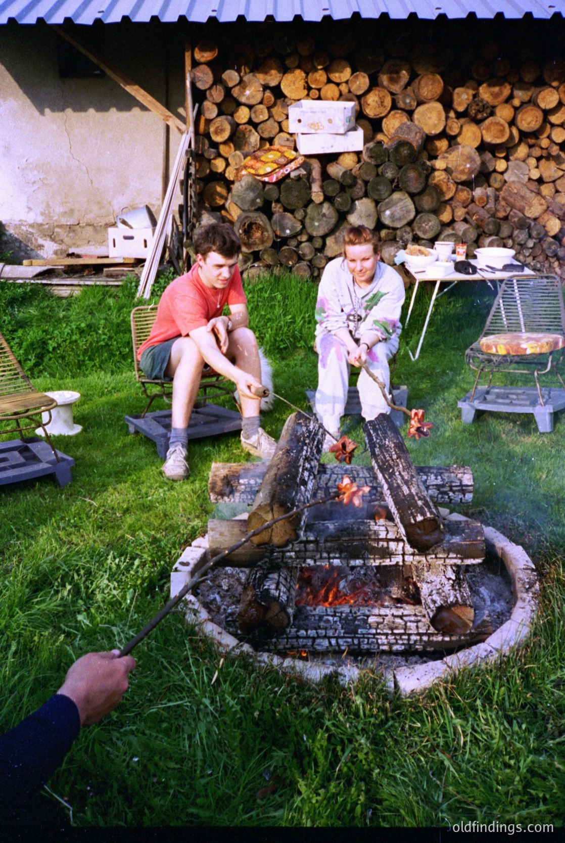 Two individuals tend a brick-built outdoor fire pit, skewering meat over flames. Stacked logs line the background wall. Mid-20th century rural or suburban setting, likely 1950s–1970s.