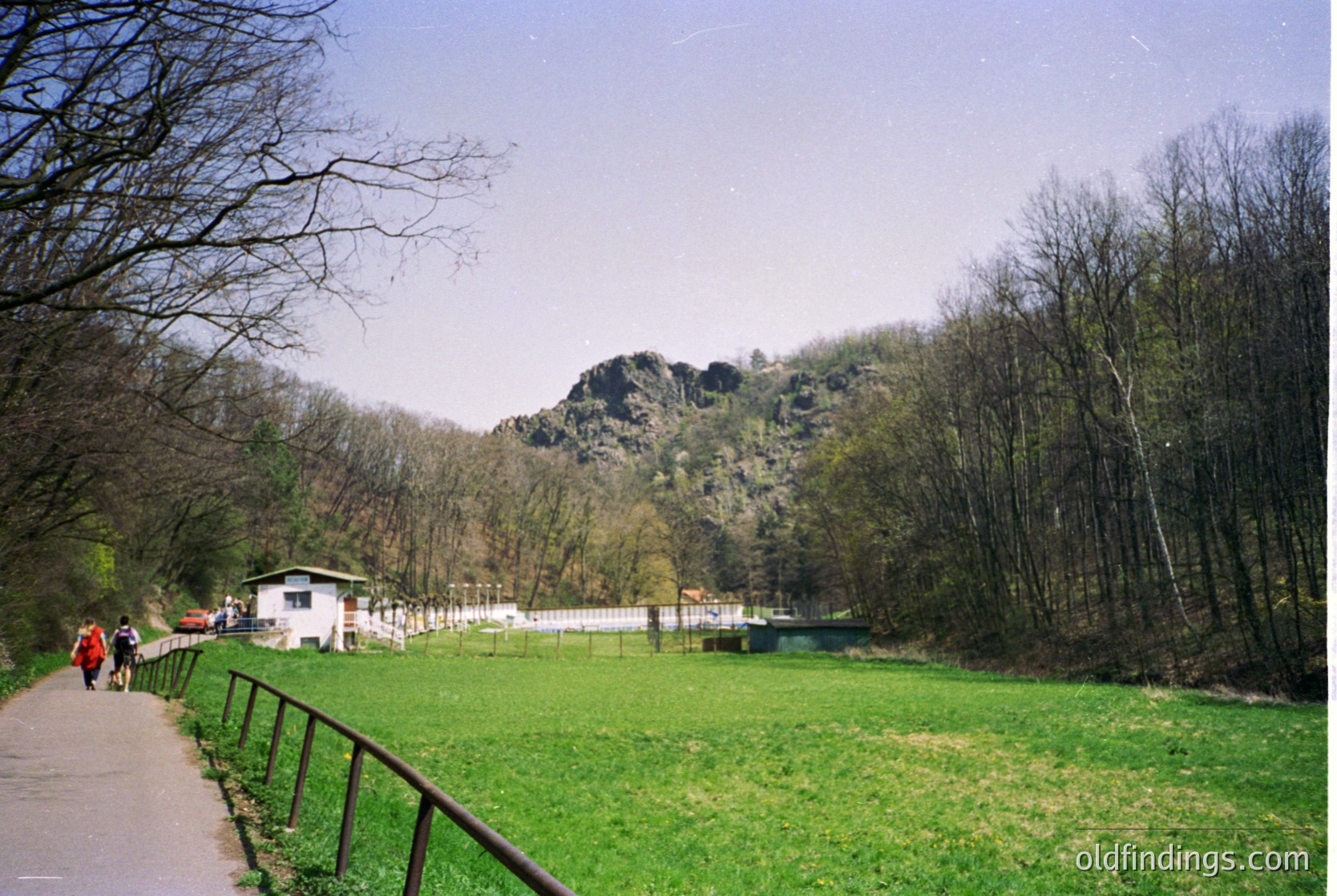 Scenic park pathway bordered by metal railings, flanked by leafless trees and lush green meadows. Prominent rocky hillside in background, with a small white building near the base. Clear skies and natural lighting suggest early spring or late autumn. Likely a public park or nature reserve.