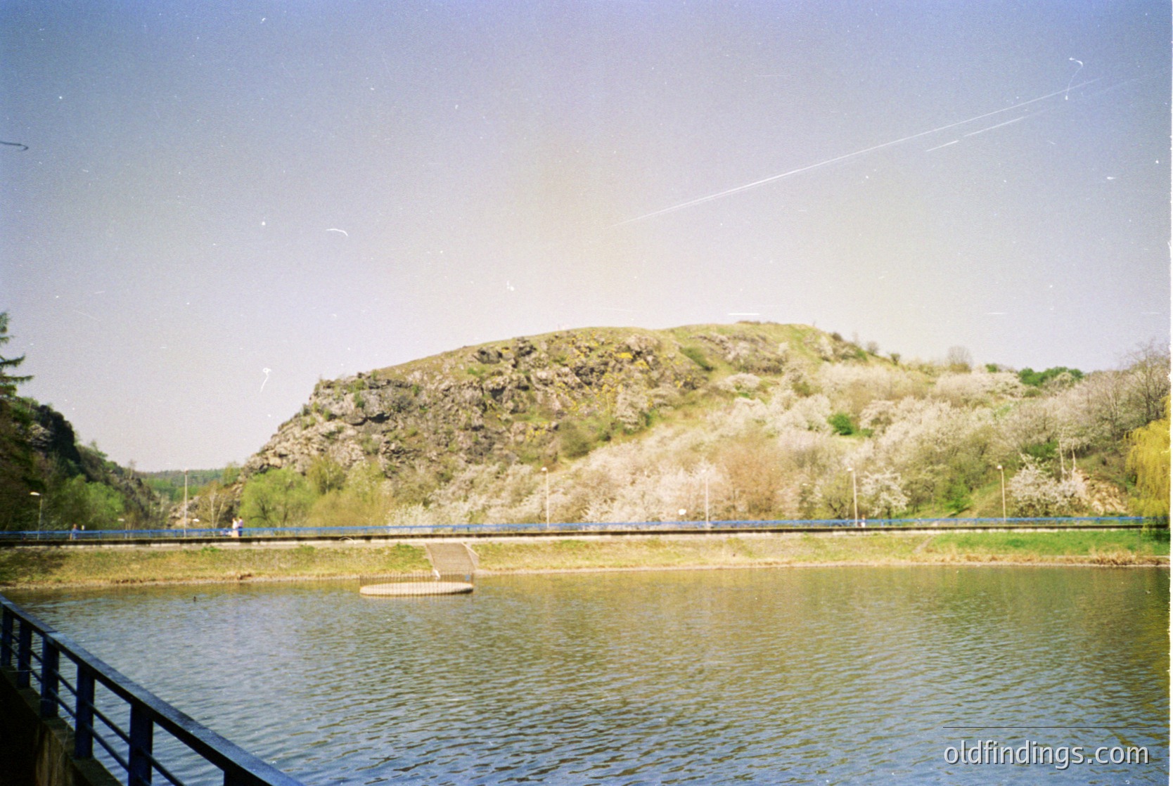 Vintage landscape shot of a serene lake bordered by a wooden walkway. Gentle slope of grassy hill in background with sparse trees. Clear water reflects muted sunlight. Likely mid-20th century due to film grain and framing.