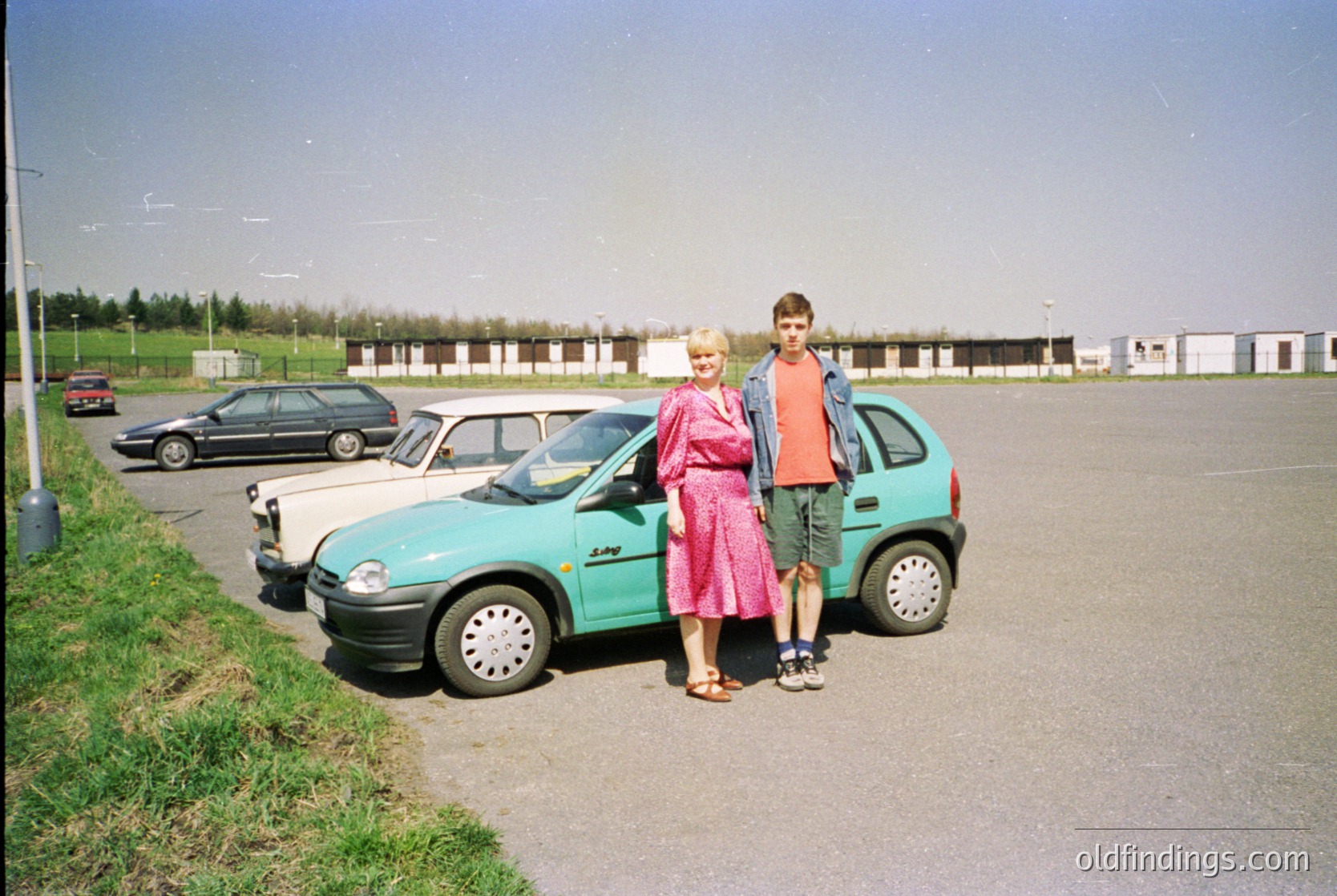 Two individuals pose beside a turquoise hatchback car in a parking lot, likely mid-1990s. Woman wears a pink blouse and floral skirt; man in a red shirt and khaki shorts. Surrounding cars suggest a suburban or small-town setting. Vague industrial buildings in background.