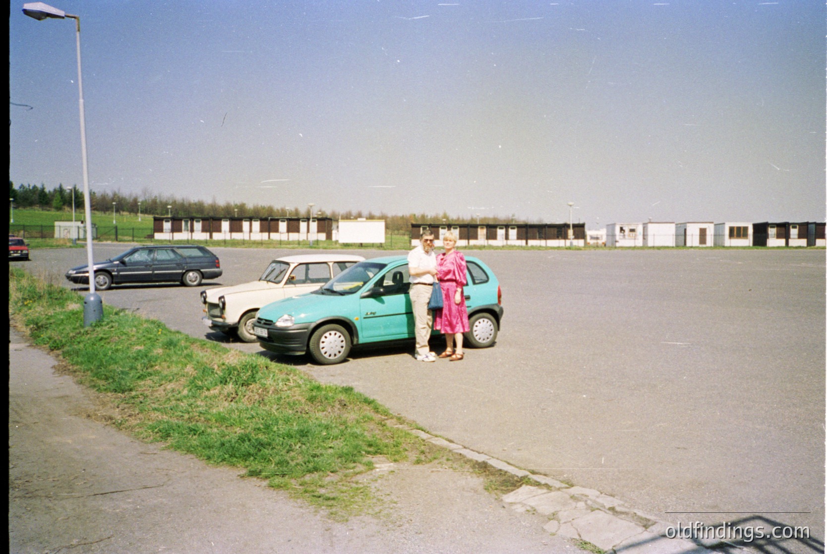 Vintage 1990s-era parking lot with two women near a turquoise hatchback. One wears a pink dress, the other a striped top. White utility buildings and parked cars (including a black sedan) in background. Grass patch along curb.