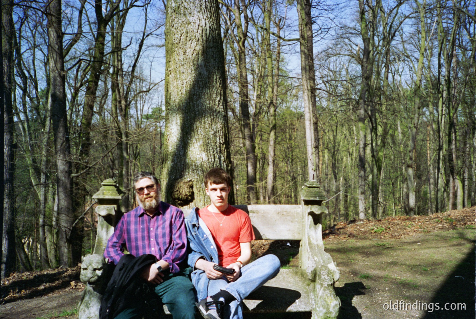 Two men pose on a weathered stone bench in a forested cemetery, surrounded by bare trees and gravestones. The man on left wears a plaid shirt and glasses; the man on right wears a red shirt and jeans. Likely late 20th century, possibly 1980s–1990s.