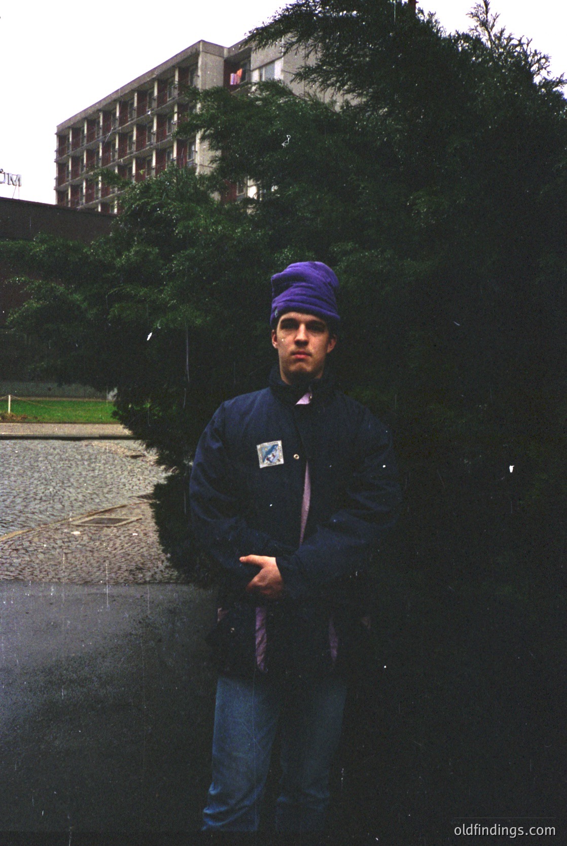 Portrait of a person in a blue raincoat and purple knit cap, standing outdoors in wet conditions. The background features a Soviet-era concrete building with a sign in Cyrillic script. Likely Eastern Europe, 1970s–1980s.