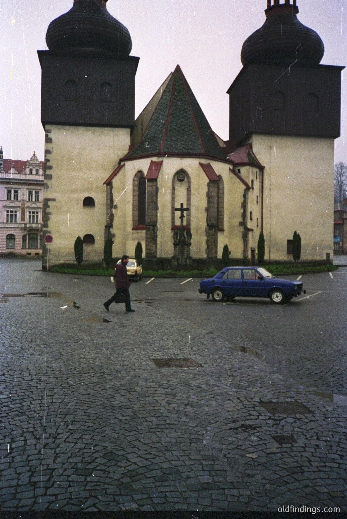 Historic Eastern European church with twin black towers and red-tiled roof, likely 1960s–70s. Cobblestone plaza features a parked blue sedan and a man in dark coat carrying a bag. Architectural details include arched windows and a central cross.