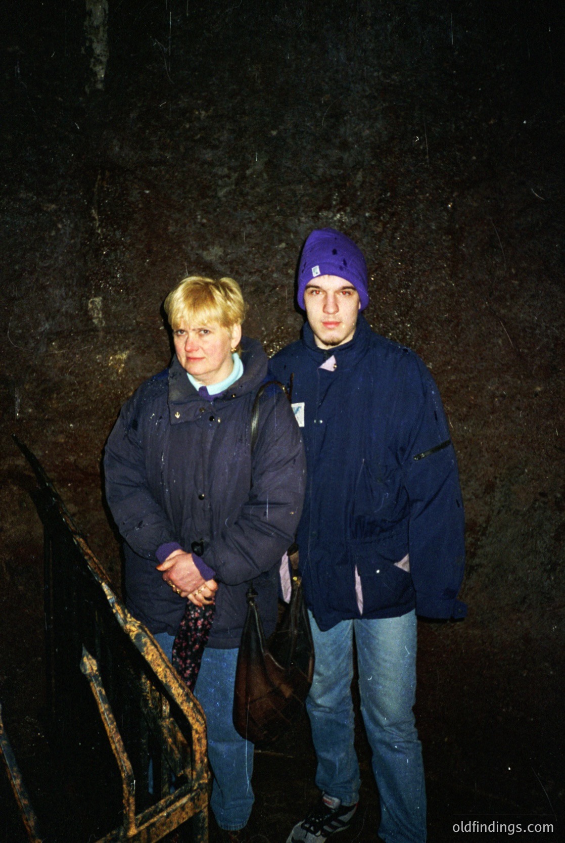 Two individuals pose indoors beside a weathered stone wall, likely part of a historic or industrial site. The man wears a navy puffer jacket with a purple knit cap, while the woman wears a matching jacket with a light scarf. Their attire suggests late 20th-century fashion (, ). The rustic wooden chair and rough stone backdrop hint at a working-class or heritage setting (, ).