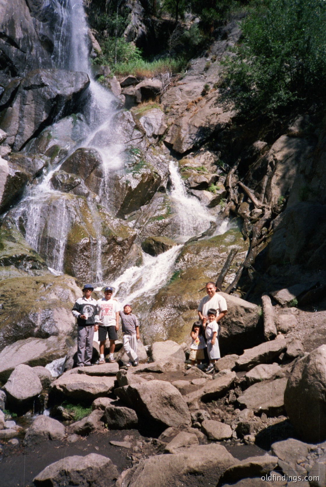 Group of 10 posing near a cascading waterfall in a rocky canyon, likely mid-20th century outdoor adventure. Casual attire suggests a family or hiking group. Lush greenery and mist enhance the alpine scenery.