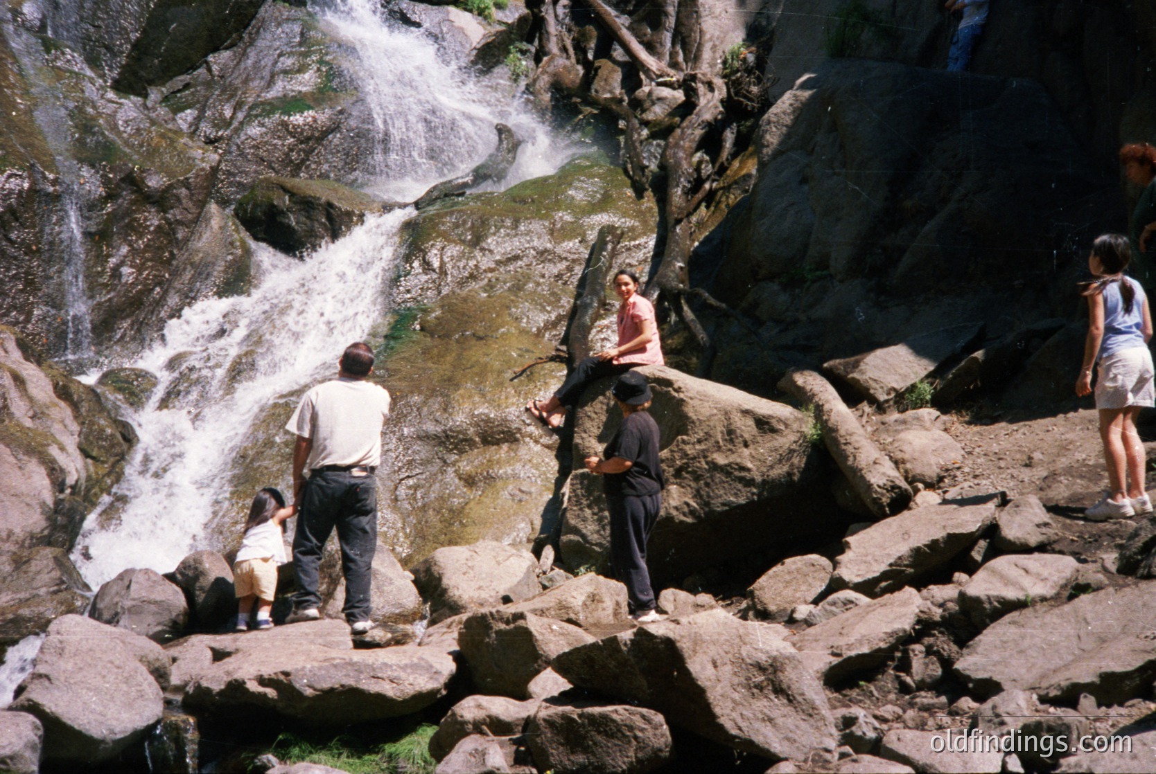 Group of five people exploring a rocky riverside waterfall trail, likely mid-1990s. Casual summer attire suggests leisure activity. Water cascades over moss-covered rocks in a narrow canyon.