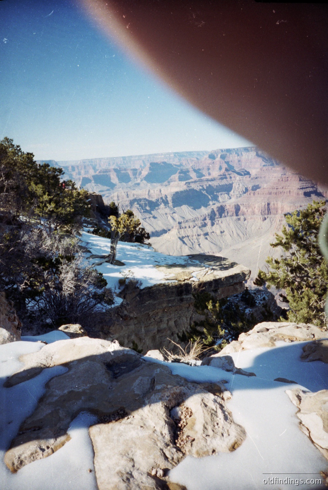 Grand Canyon’s layered red rock formations under clear skies, captured from an elevated viewpoint. Snow patches highlight winter contrast. Pine trees frame the edge, emphasizing scale. Dramatic lighting and perspective evoke natural grandeur.