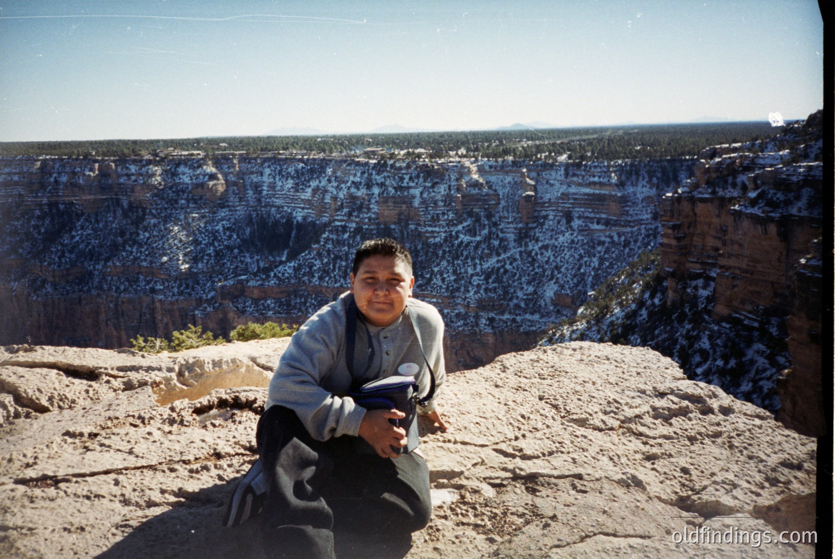Person kneeling on Grand Canyon rim, holding a camera, dressed in 1990s-style fleece and dark pants. Striking layered red rock formations and distant pine forests below. Clear blue sky with minimal clouds.