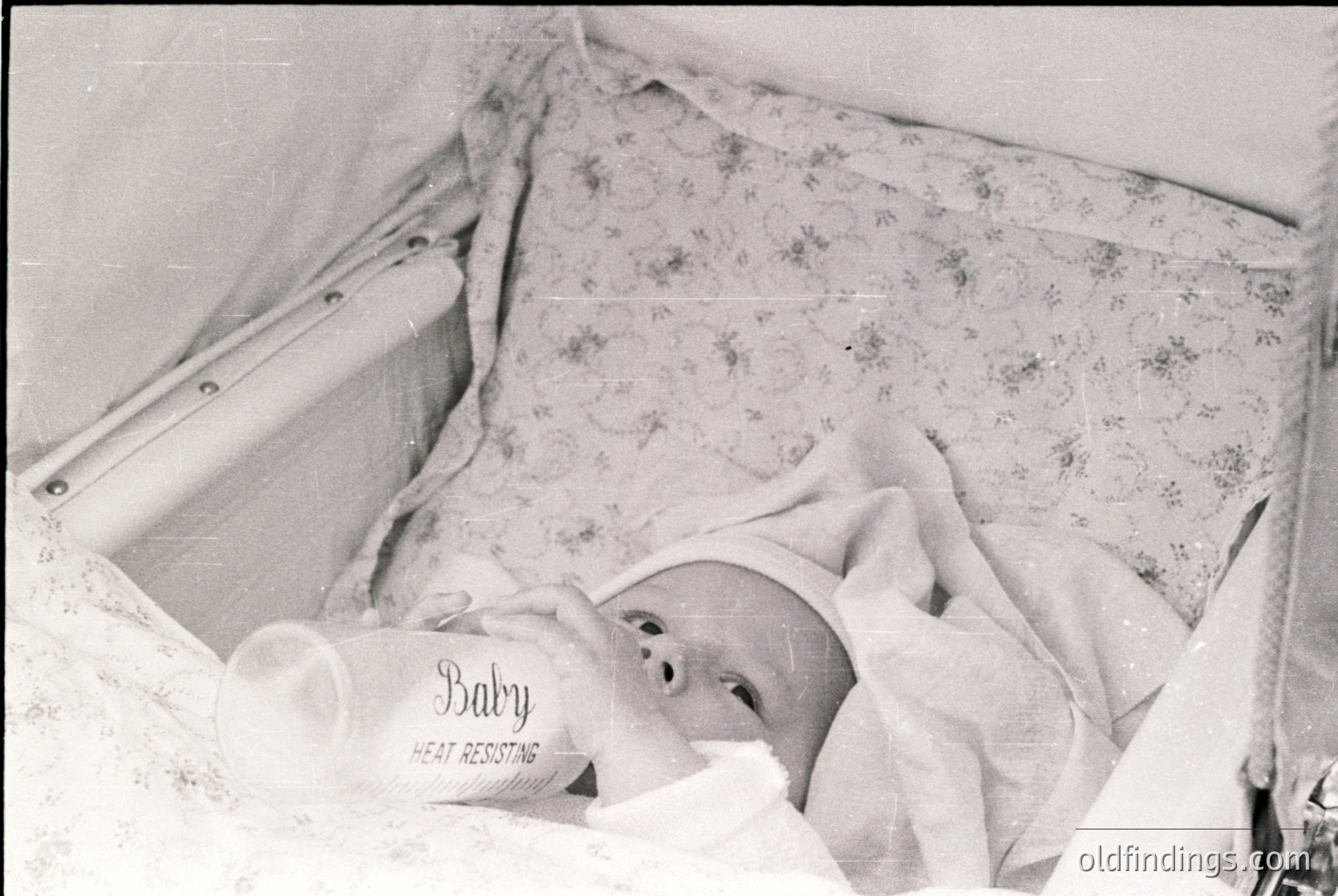 Vintage black-and-white photo of an infant in a **Baby Heat Resisting** blanket, likely a 1950s–1960s medical or hospital setting. The infant lies in a crib with floral-patterned bedding. Medical context suggests neonatal care or pediatric ward.