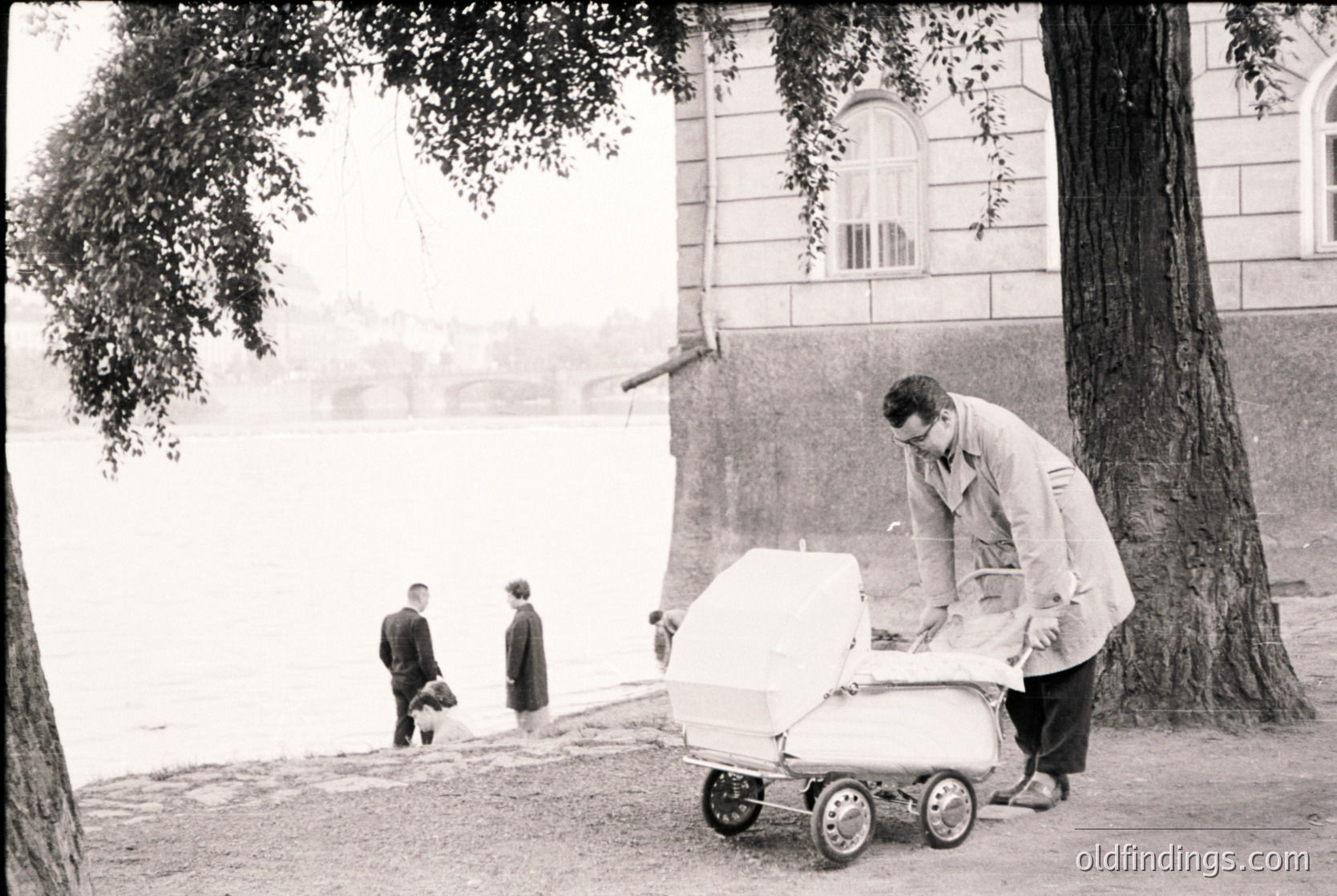 Mid-20th century black-and-white street scene featuring a man in a light coat pushing a vintage baby stroller near a grand, classical-style building with arched windows. Three children in dark clothing walk in the background along a tree-lined path. Urban park or residential area, likely post-WWII Europe.