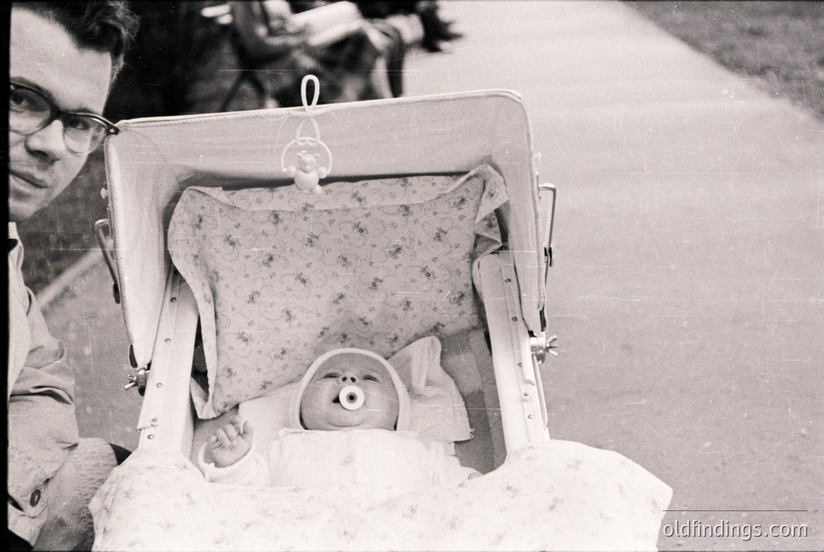 A man in mid-20th-century attire holds an open suitcase repurposed as a portable bassinet, containing a sleeping infant wrapped in a patterned blanket. The scene suggests travel with a child, likely or .