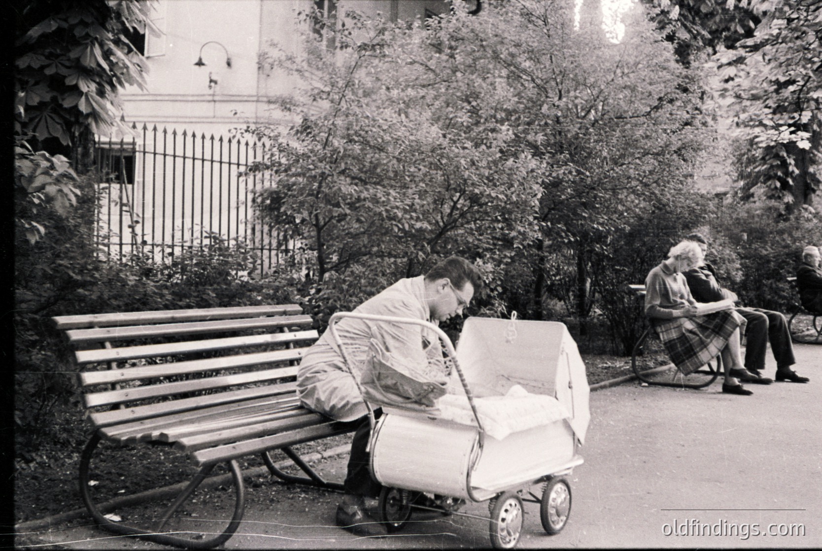 Mid-20th century urban park scene: man in light jacket adjusts vintage stroller beside wooden bench. Elderly woman in plaid sits nearby, reading. Iron fence and leafy trees frame the composition. Likely Eastern European setting, 1950s-1960s.
