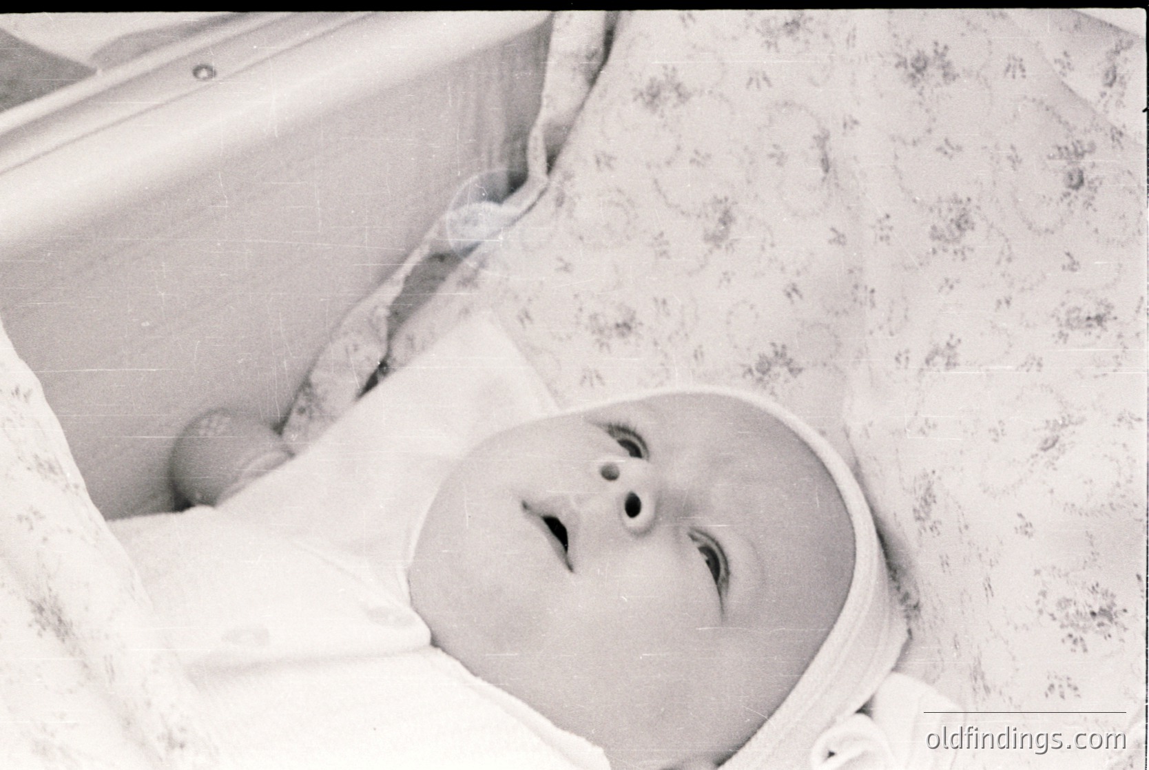 Vintage black-and-white portrait of an infant in a crib with floral-patterned linen, mid-20th century. Classic baby cap and diaper visible. Evokes nostalgic family archives or historical parenting trends.