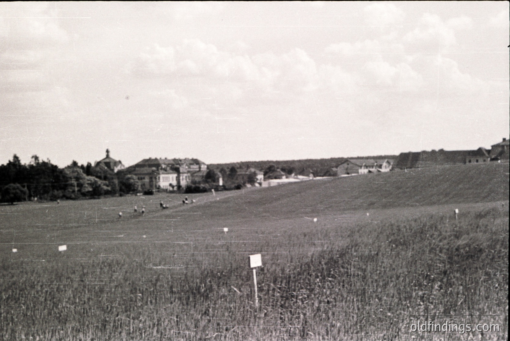 Mid-20th century rural landscape with scattered farmhouses and open fields, likely Eastern Europe. Black-and-white photo captures low-lying buildings, grazing livestock, and a signpost in foreground.