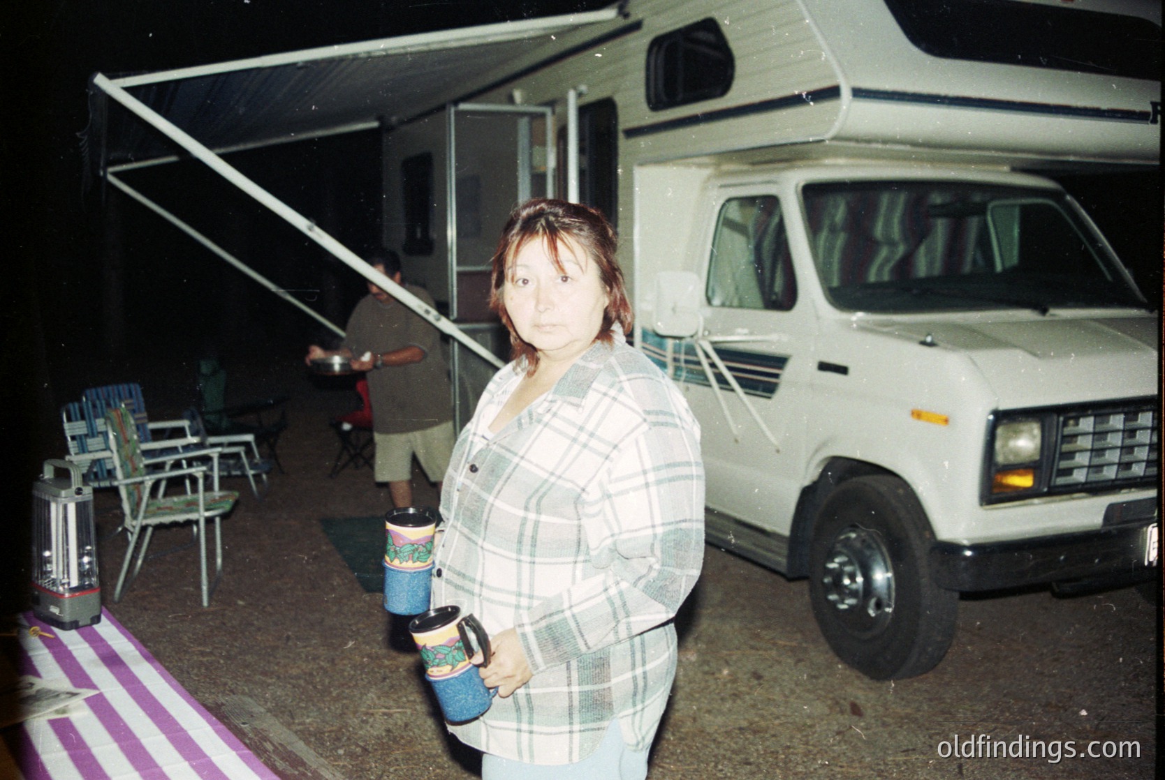 Vintage camper van parked under a canopy at night, featuring classic 1980s design with pop-up roof. Woman in plaid shirt holds two disposable coffee cups. Foldable chairs and a portable grill visible in background. Likely a road trip or camping scene.