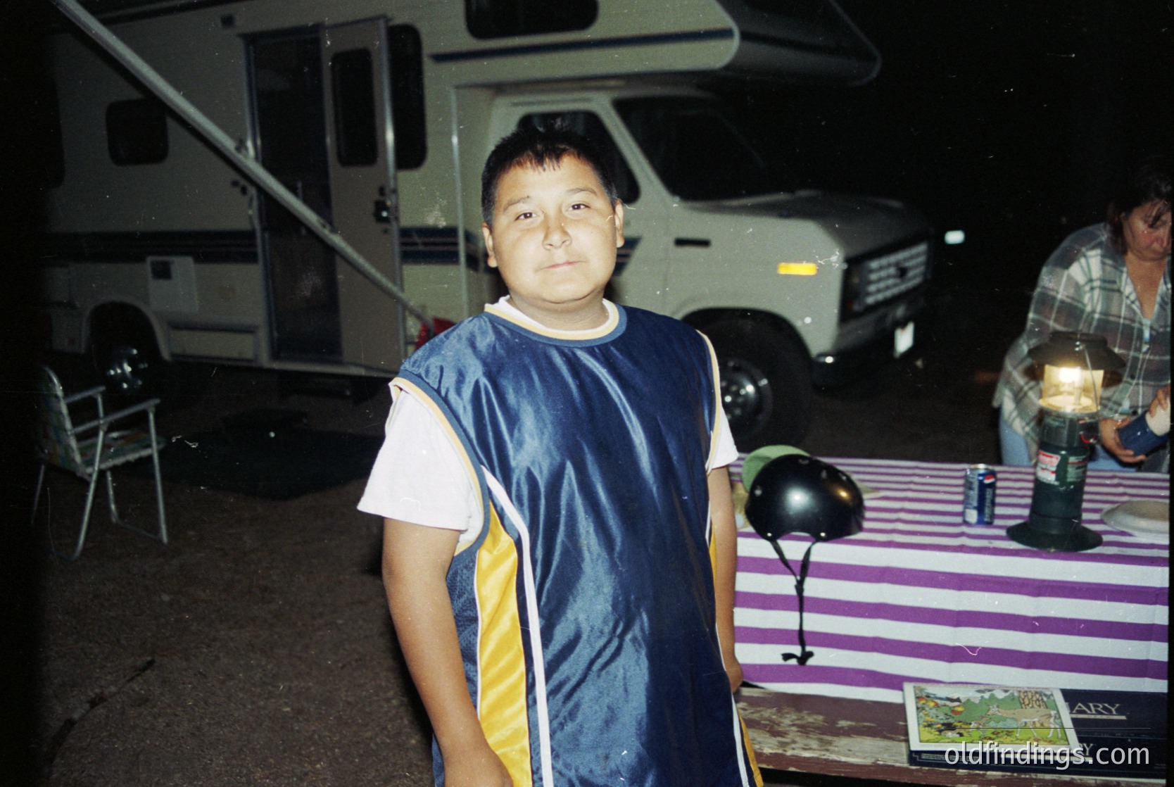 Vintage RV campground scene featuring a young boy in a navy-blue sleeveless shirt with yellow stripes, standing beside a striped tablecloth. White trailer and lantern-lit table with camping gear in background. Likely late 20th century, possibly or . Evokes nostalgia for outdoor family trips.