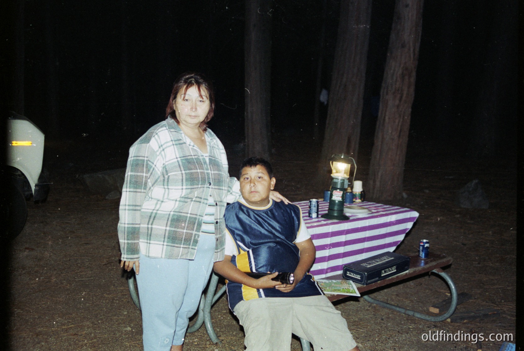 Vintage candid shot of two individuals posing outdoors at night, likely 1990s–2000s. Woman in plaid shirt and light jeans stands beside a seated boy in a dark polo and khaki shorts. Rustic wooden table with striped tablecloth holds a lantern, soda cans, and a cooler. Dark, natural setting suggests rural or backyard gathering.