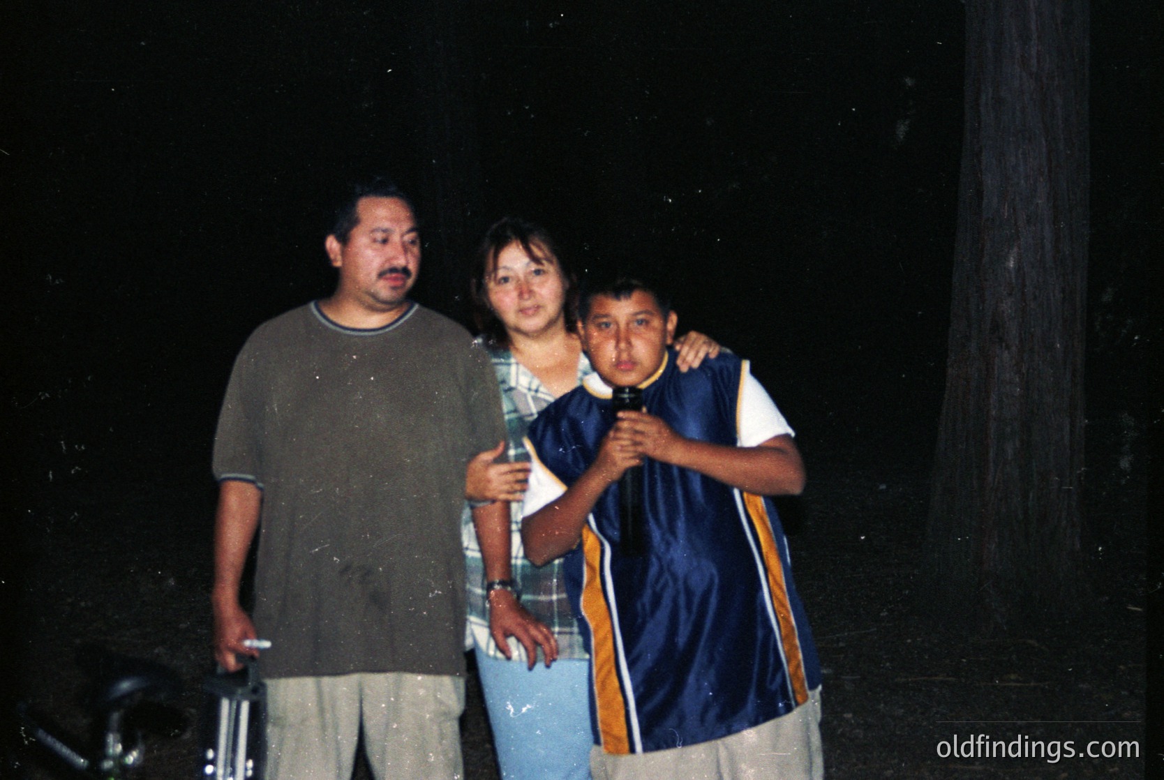 Three individuals pose outdoors at night under dim lighting, likely flashlit. The man wears a gray T-shirt and beige pants; the woman, a checkered shirt and light blue pants; the boy, a blue hoodie with gold stripes and light shorts. Their expressions appear candid. Background suggests a rural or semi-rural setting with wooden structures.