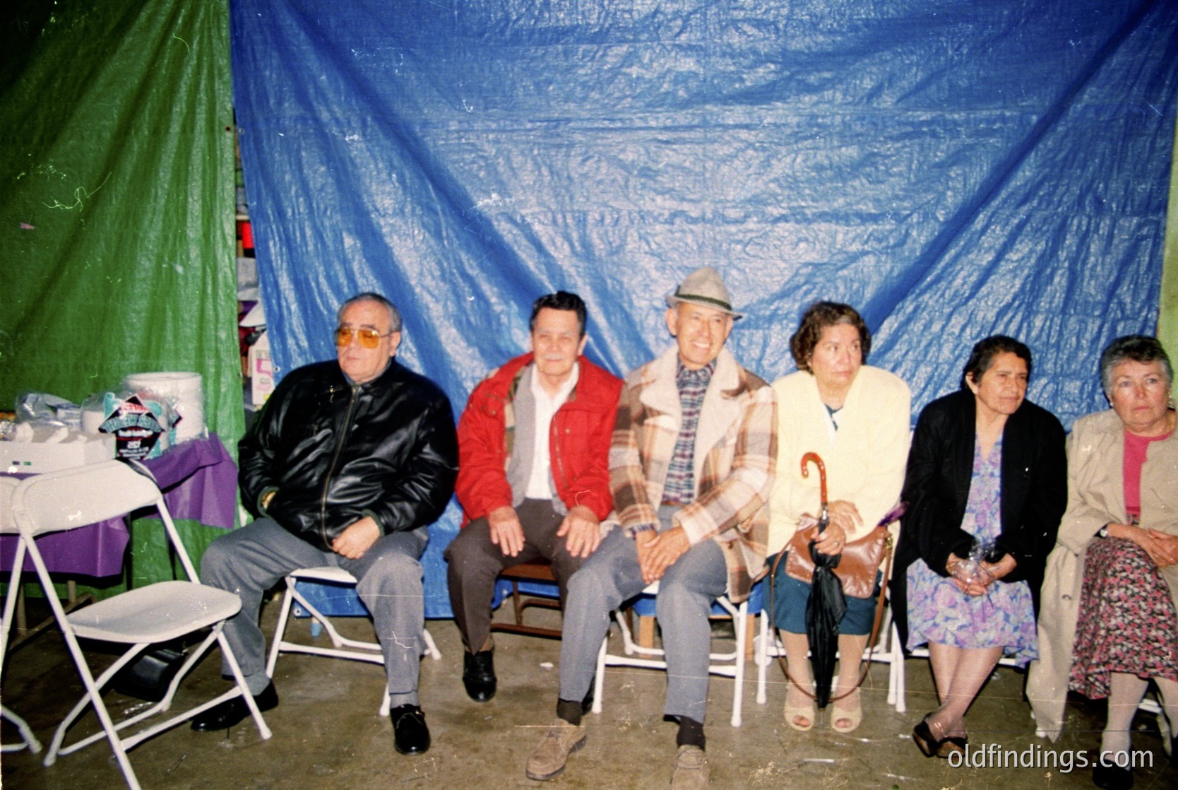 Five adults pose indoors under blue tarpaulin, seated on metal chairs. Men wear jackets (one in red plaid), hats, and glasses; women in floral dresses and one with an umbrella. Faded lighting suggests vintage setting. Likely 1970s–1990s, informal gathering or event.