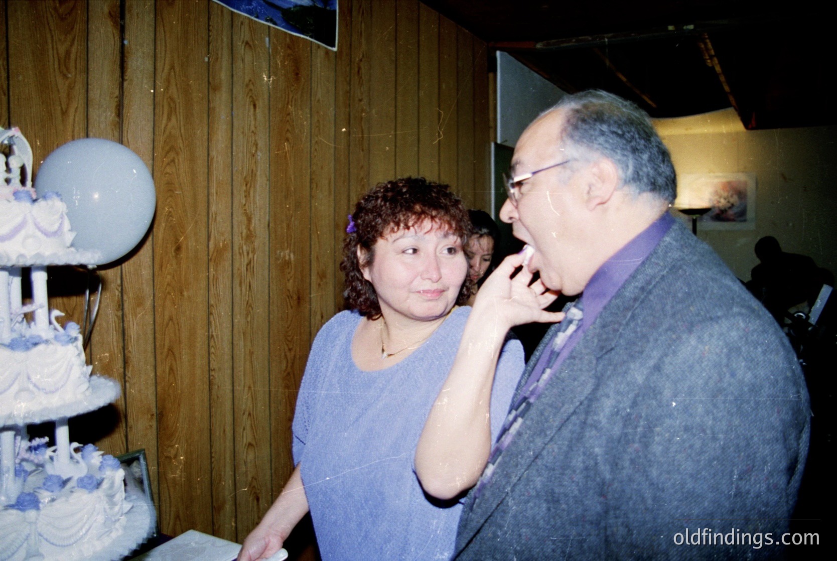 Vintage indoor celebration featuring a multi-tiered wedding cake with floral decorations. A woman in a purple dress and man in a grey sweater share a moment, possibly toasting or whispering. Warm lighting and wooden paneling suggest a cozy, intimate venue. Likely late 20th century (1980s–1990s).
