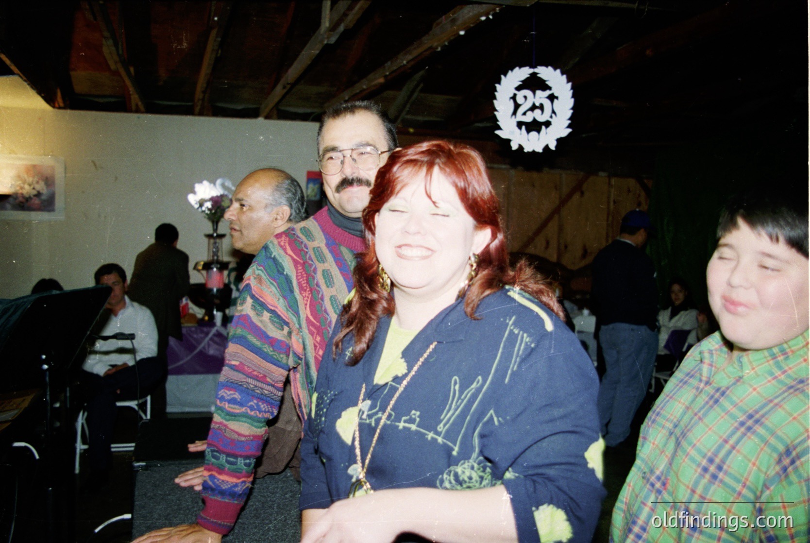 Vintage indoor gathering featuring three smiling adults in casual 1990s attire—plaid sweater, graphic tee, and checkered shirt. Warm lighting and a festive logo (resembling a crown) on a draped backdrop suggest a themed event or celebration. Background includes blurred attendees and a partially visible mural.