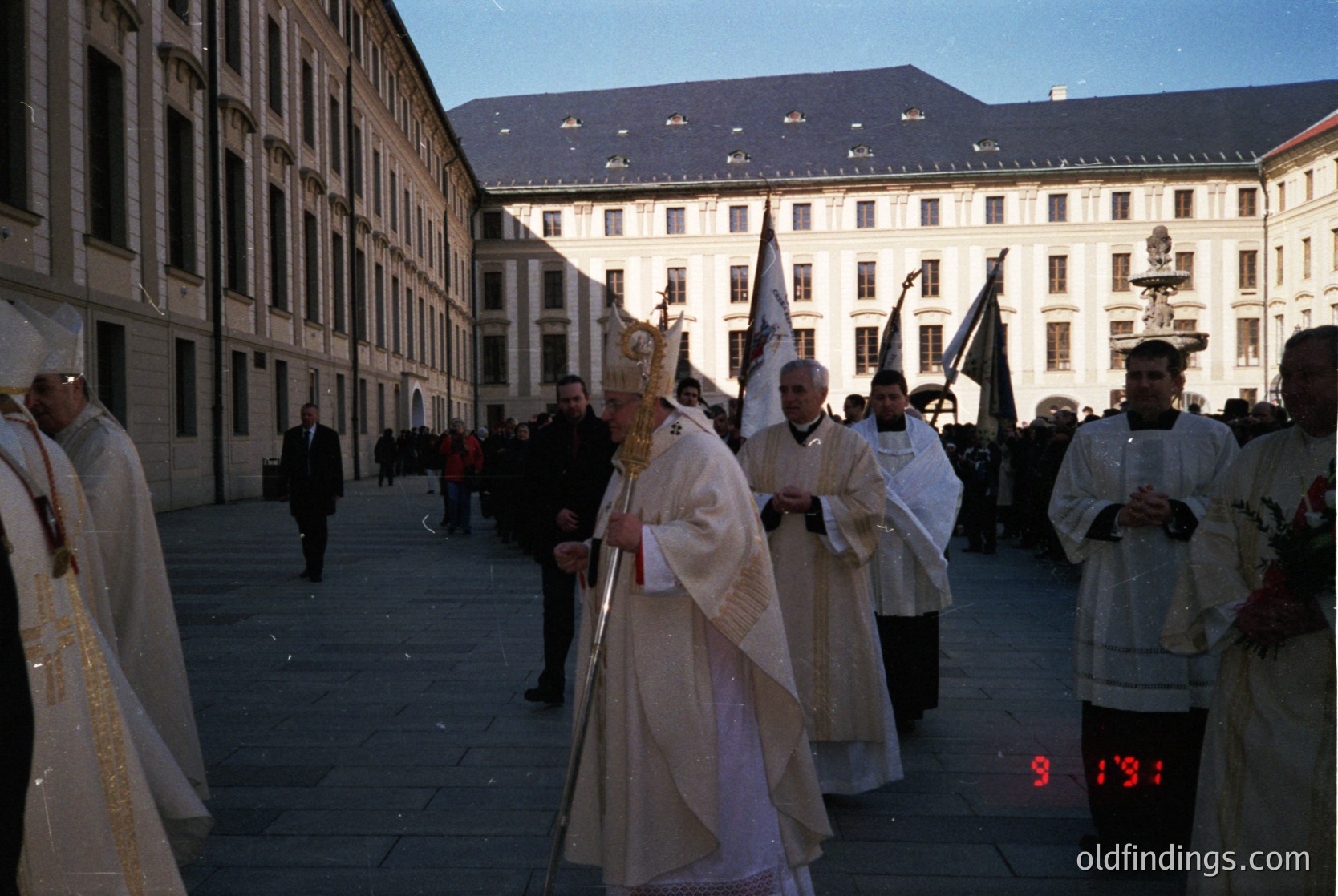 Religious procession in a European courtyard, likely 1970s–1980s. Clergy in ornate liturgical vestments, including a mitre and crosier, lead a solemn march. Architectural backdrop features classical stone buildings with arched windows. Crowd gathers along the perimeter, observing.