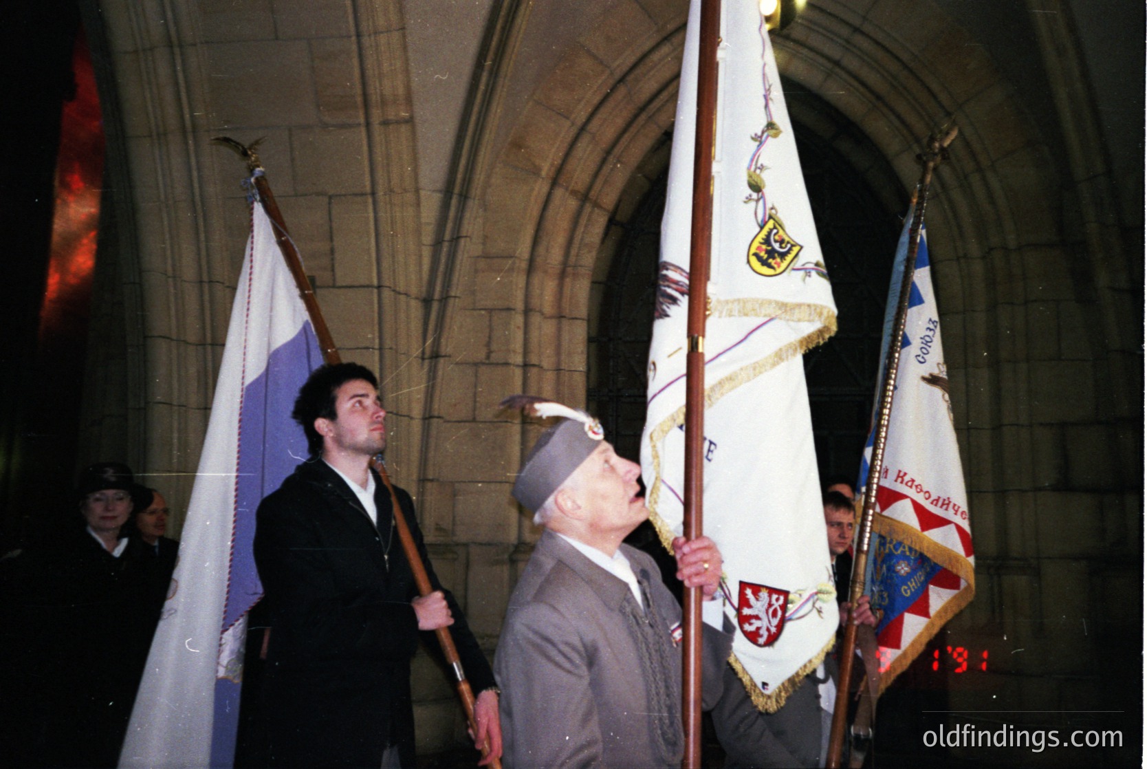 Veterans in formal attire holding flags under a stone archway, likely at a WWII commemoration. The central figure wears a 1930s-40s military cap and a dark suit, holding a flag with Cyrillic text and a 1934 emblem. Another flag features a blue, white, and red striped design. Gothic-style architecture suggests a European setting, possibly Eastern Bloc.