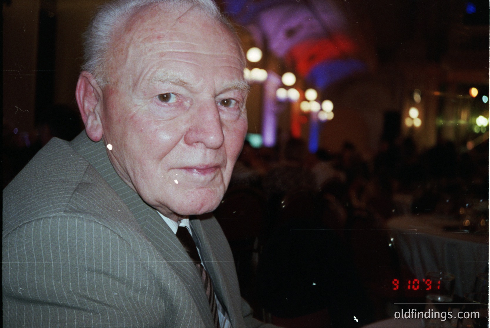 Portrait of an elderly man in a formal suit, captured in low-light indoor setting. His expression is neutral, with a slight smile. The blurred background suggests a social event with festive lighting and a digital clock displaying "3:10." [Mid-20th century formal portrait, vintage photography, evening event, classic men's attire, social gathering]