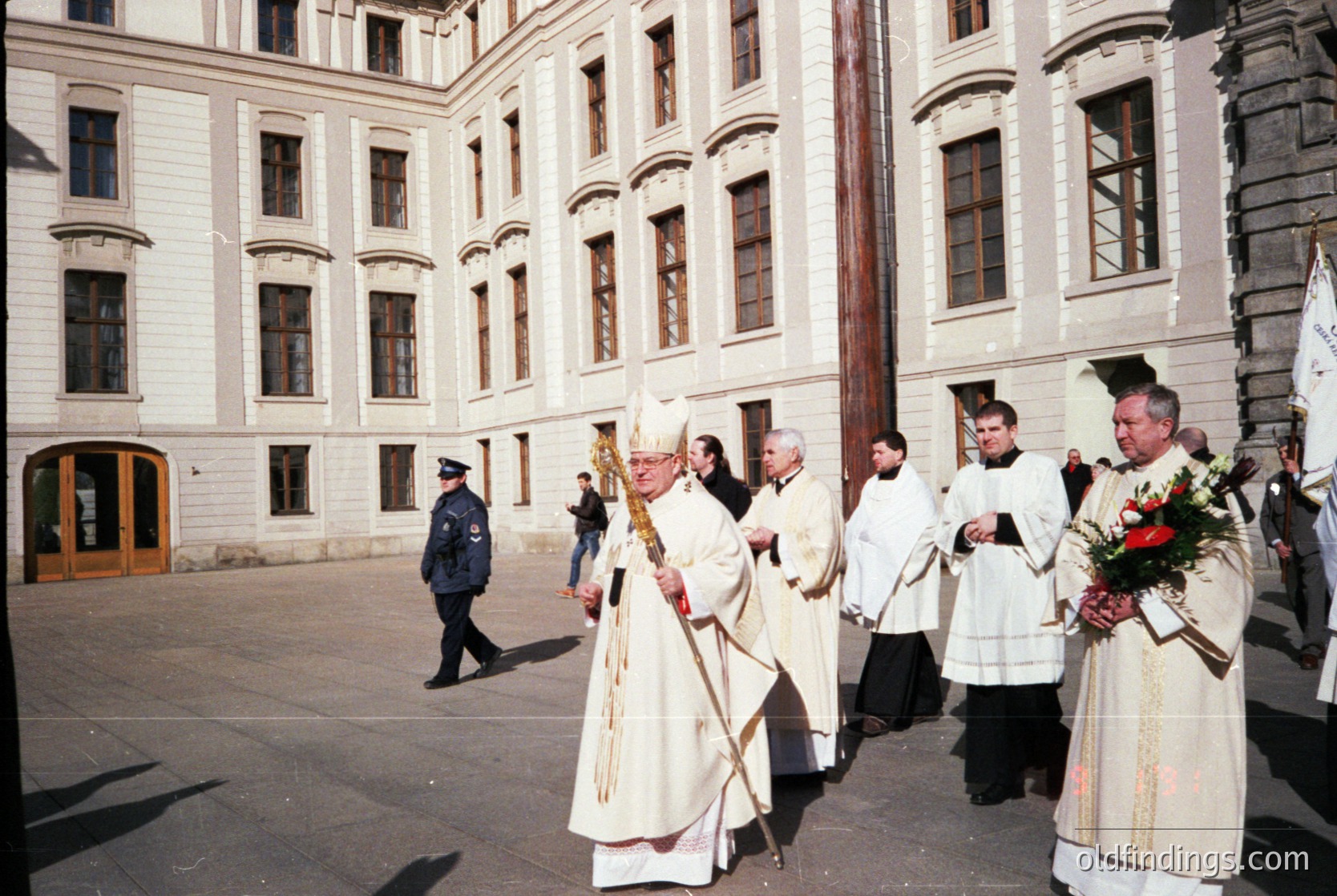 Religious procession in a courtyard featuring neoclassical architecture. Clergy in liturgical vestments, including a bishop with a mitre and crosier, lead the ceremony. Uniformed officers in the background suggest official presence. Likely Eastern Orthodox given vestments and setting.