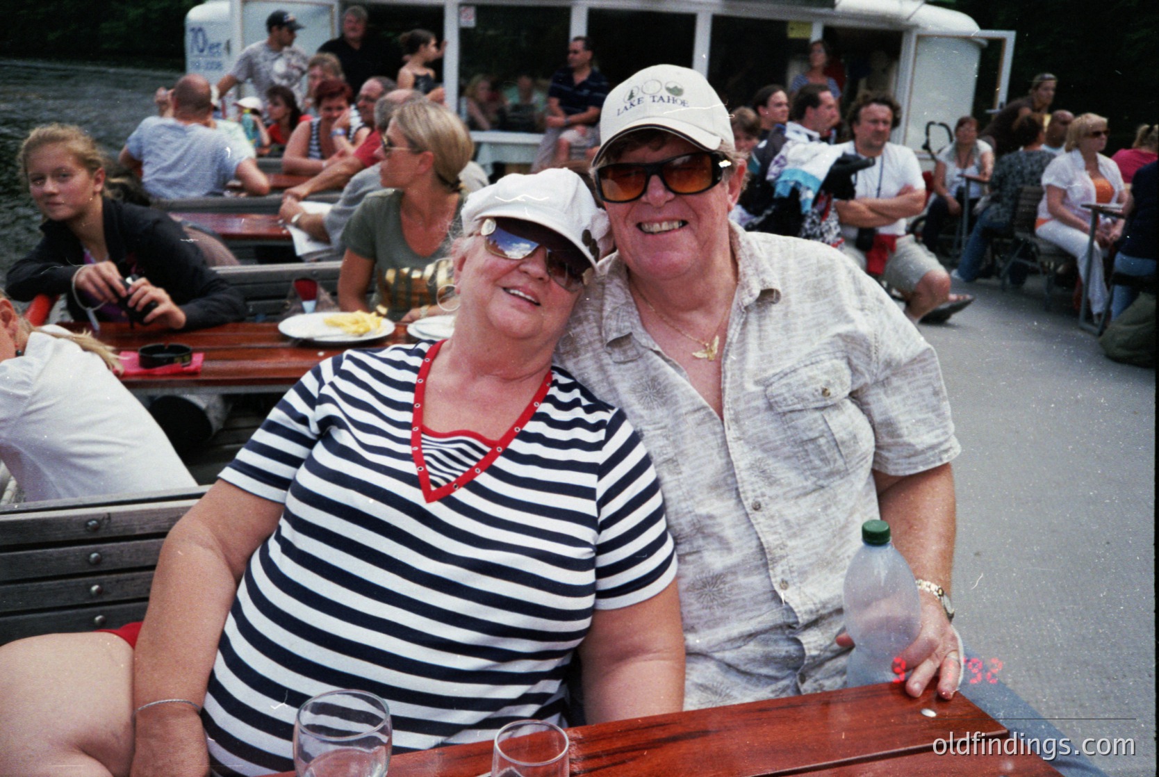 Retro riverboat scene with two smiling adults in 1980s summer attire—woman in striped tank top, man in cap and sunglasses. Crowded wooden benches filled with casual diners. Possible European canal or waterfront setting.