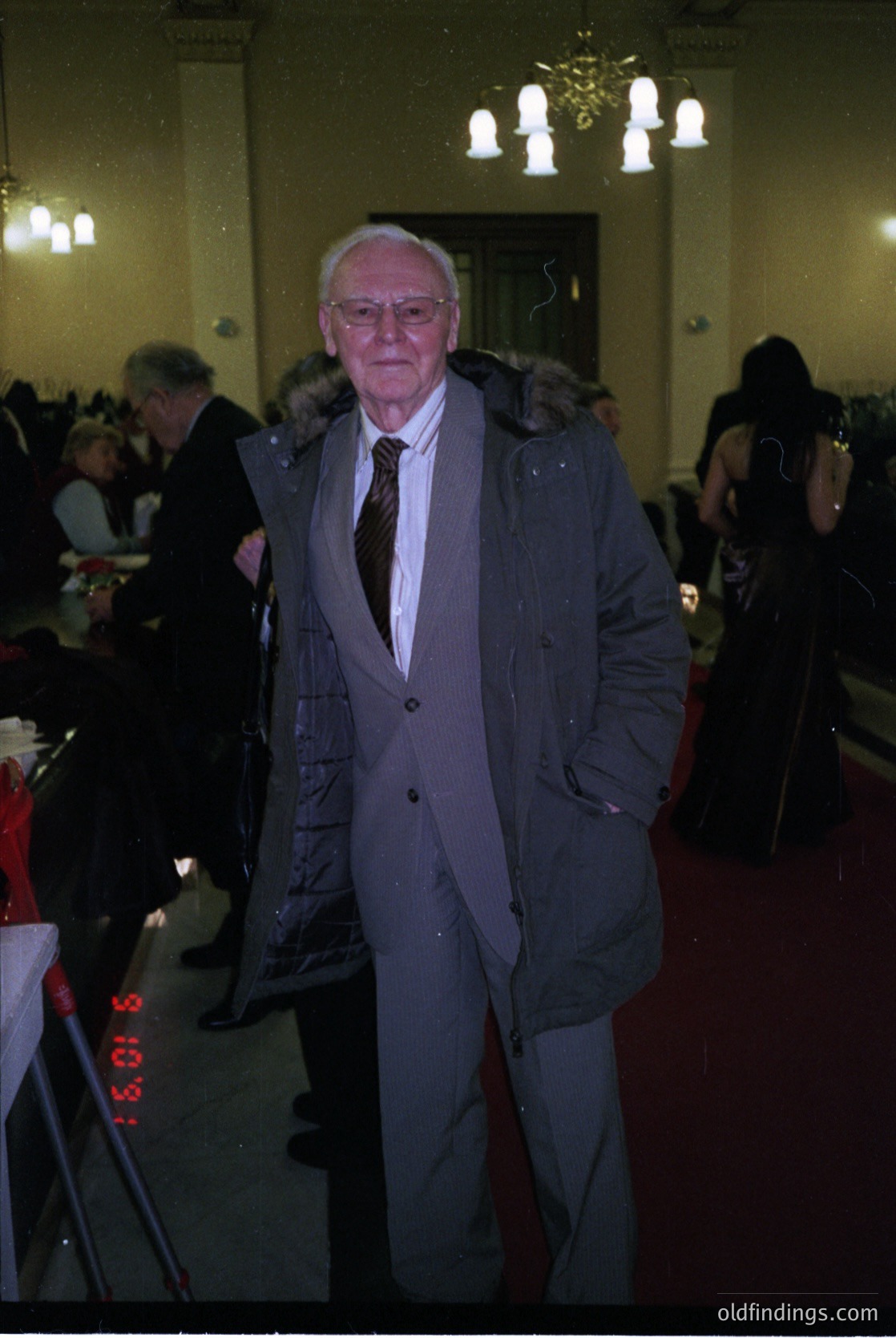Mid-century formal event in a grand hall, featuring a man in a dark blazer, striped tie, and glasses. Ornate chandeliers and vintage wall sconces illuminate the space. Blurred attendees suggest a lively gathering, possibly a wedding or banquet. Style reflects 1960s–1970s elegance.