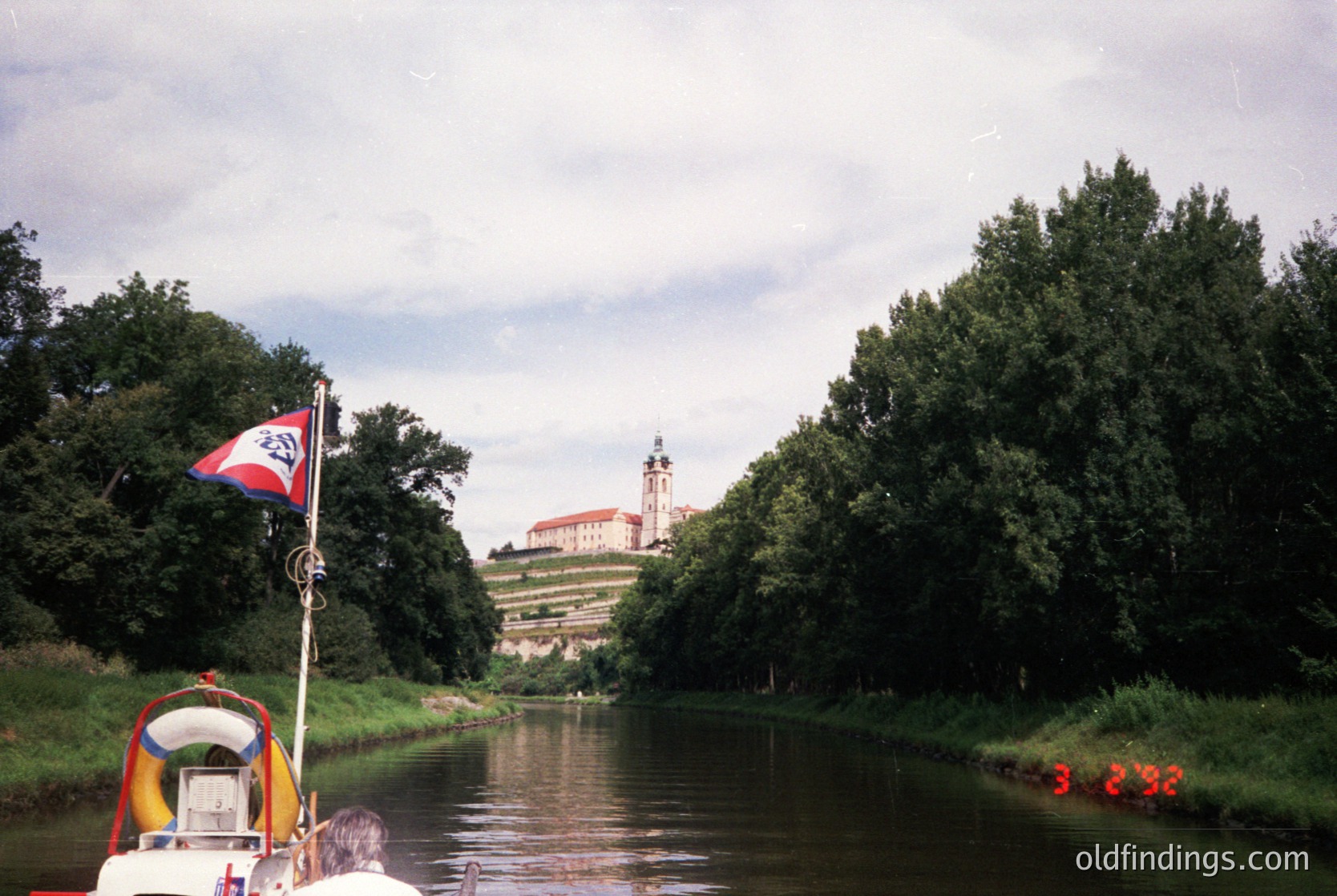 Historic canal scene with a flag bearing a white emblem on red/blue field, likely . A boat with red life vests cruises past lush greenery, leading to a castle-like structure with a prominent tower on a hill. --- *Notes: Flag resembles the Czech coat of arms, and architectural style suggests a Central European castle. Life vests and boat design suggest mid-to-late 20th century.*
