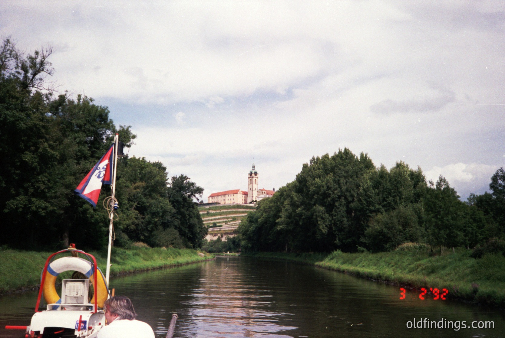 A vintage boat cruises a calm river flanked by lush greenery, with a castle-like complex in the background. The structure features a prominent tower and classical architecture, likely from the 19th century. A flag with a red and white emblem waves on a pole. [Historic riverside castle complex with boat cruise, lush greenery, 19th-century architecture ]